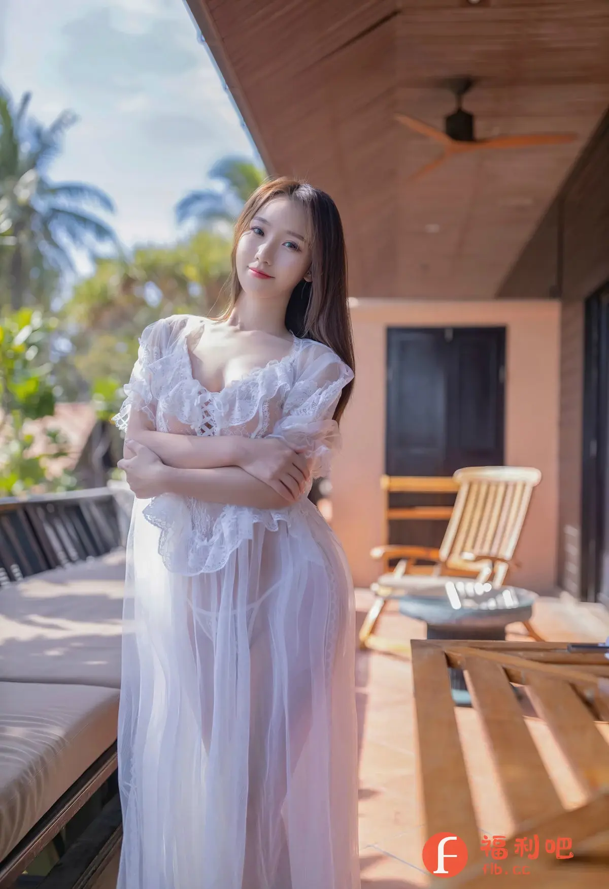 这张照片拍摄的是一位亚洲美女的私房写真，她站在一个阳光明媚的户外阳台上。背景是棕榈树和蓝天，营造出一种热带度假的氛围。
美女穿着一件轻盈的白色透明长裙，裙子带有蕾丝和褶皱的细节，显得性感而飘逸。透过裙子，可以看到她纤细的身材和内衣。她交叉双臂，眼神温柔地看向镜头，展现出自信和魅力。
阳台上摆放着木制长椅和休闲椅，增添了舒适感。天花板上有一个复古的吊扇。整体构图和光线都非常出色，突出了美女的美丽和性感，同时也营造出一种轻松愉悦的氛围。