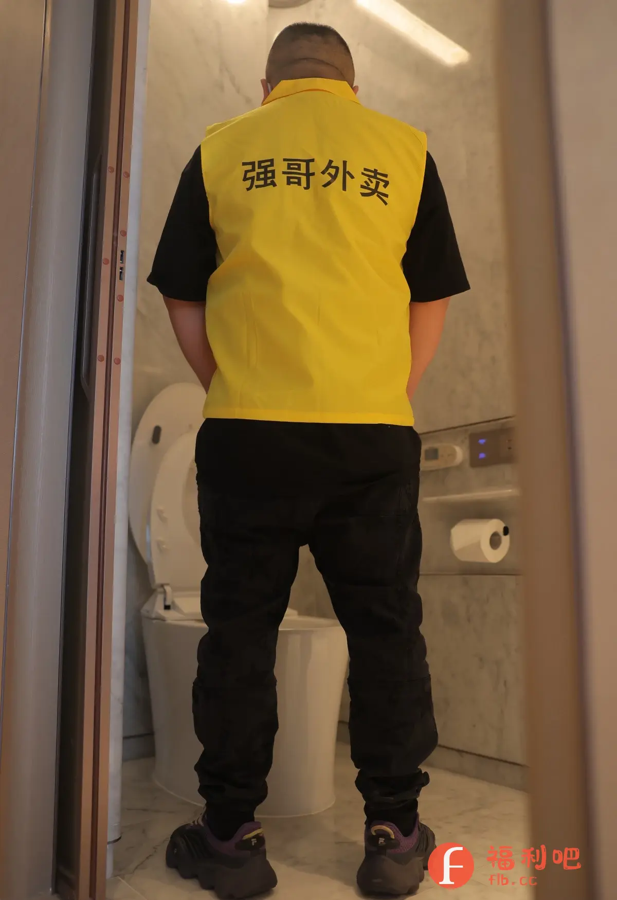 描述:一个男人穿着黑色裤子、黑色T恤和黄色马甲站在卫生间里,背对着镜头。马甲上印有汉字“强哥外卖”。他的前面是一台马桶,卫生间的墙壁是白色大理石,墙上挂着卷筒卫生纸。