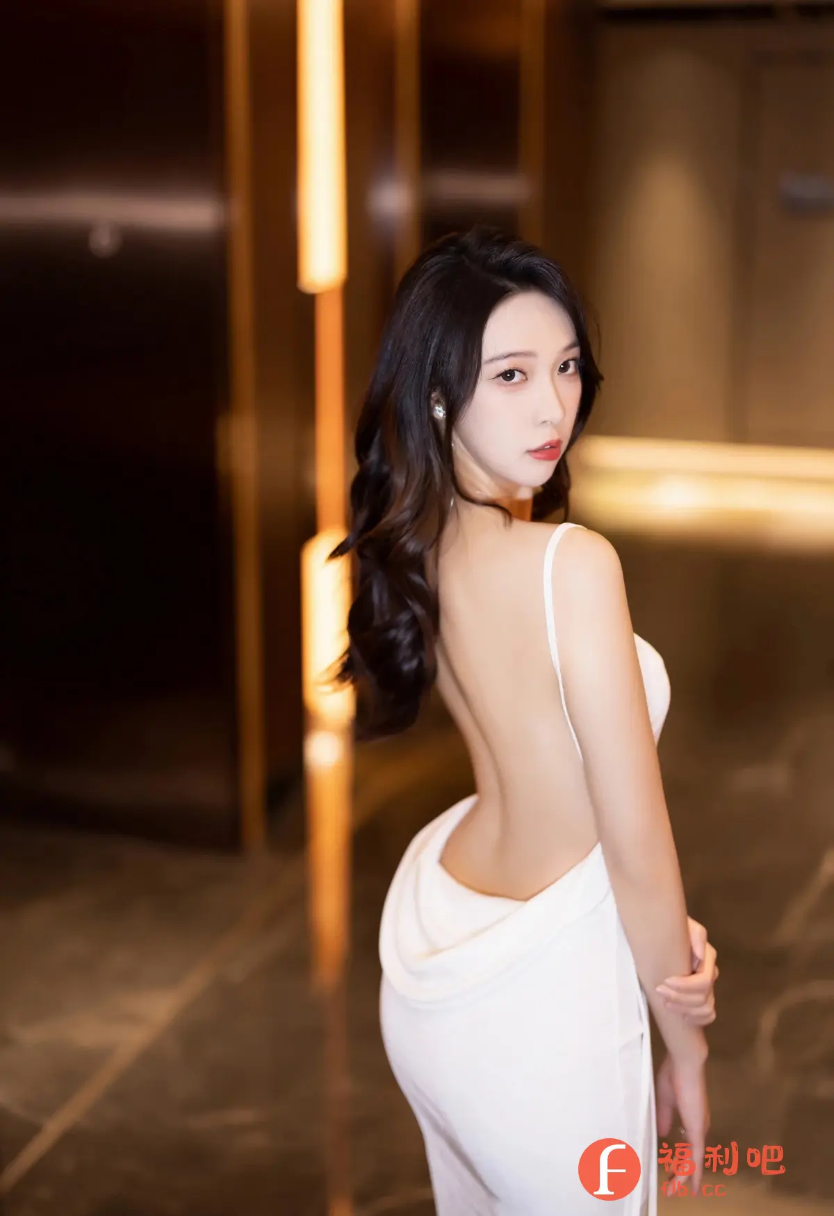 照片中是一位穿着白色露背礼服的美女。她回眸望向镜头,眼神迷人,红唇性感。她的黑色长发呈波浪状披在肩上,更显妩媚动人。礼服的设计突出了她的身材曲线,背部的线条非常优美。背景是豪华的室内环境,墙面上的灯光营造出一种温馨而浪漫的氛围。整体构图和光线运用都非常出色,展现了美女的优雅气质和迷人魅力,是一张高质量的写真作品。