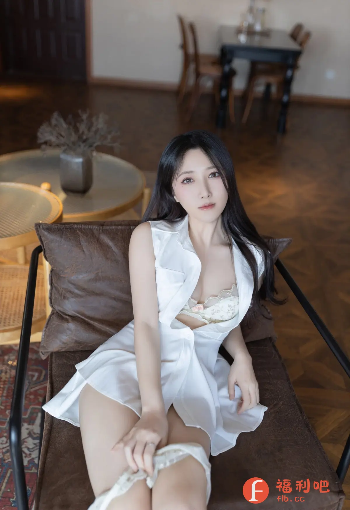 照片中,一位年轻的亚洲女性坐在棕色的皮质椅子上。她身穿一件白色无袖衬衫式连衣裙,略微敞开,露出精致的内衣。内衣是淡色的,带有精美的花纹和细节。她的长发乌黑亮丽,自然地垂在肩上,衬托出白皙的肌肤。她的眼神柔和,表情平静,略带一丝神秘感。她的手轻轻地放在腿上,指间戴着一枚戒指。整体光线柔和,营造出一种温馨而私密的氛围。背景是室内环境,可以看到一些家具,如圆形桌子和装饰品,但都被虚化处理,突出了人物主体。