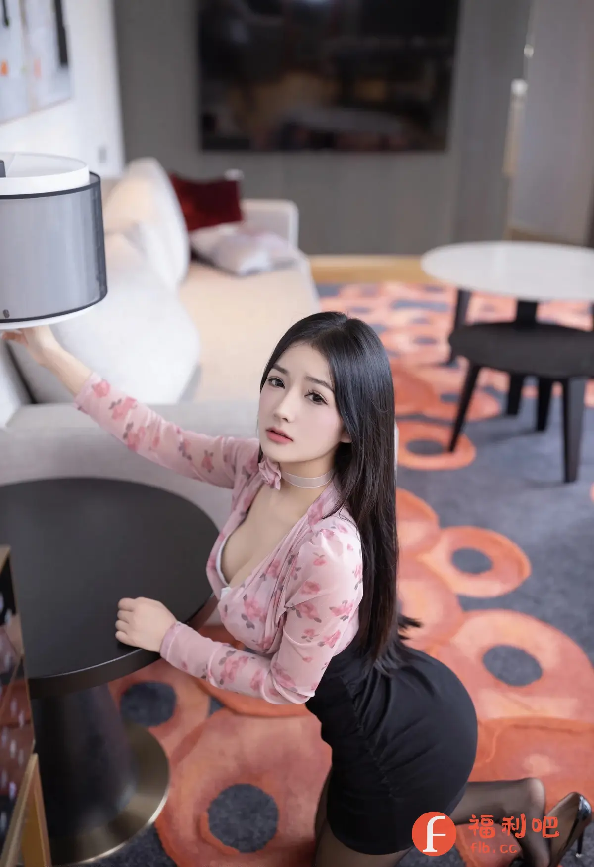 照片中，一位年轻女性跪在地板上，她的姿势和衣着透露出一种挑逗的氛围。她穿着一件带有粉色花朵图案的透明上衣，里面隐约可见白色的内衣，下身则是一条紧身的黑色短裙，搭配着黑色的丝袜和高跟鞋。她的长发垂直地垂在肩上，眼神直视前方，略带一丝妩媚。她一只手搭在黑色的圆形桌子上，另一只手则似乎在调整旁边的灯罩。
背景是一个装饰现代的房间，可以看到沙发、茶几和带有抽象图案的地毯，整个场景给人一种私密和个人的感觉，暗示着这可能是在一个酒店房间或者公寓里拍摄的。