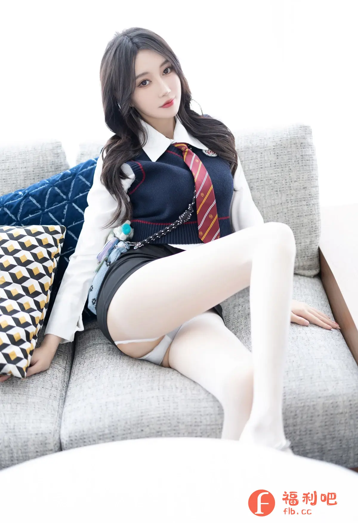 照片中一位年轻女性穿着校服风格的服装,展现出青春活力的形象。她身穿一件白色衬衫,外面搭配一件深蓝色针织背心,配上一条带有红色的条纹领带,胸前还佩戴着一枚徽章,整体造型充满了学院风。下身穿着一条黑色短裙,搭配白色丝袜,更显腿部线条的修长。她随意地坐在灰色的沙发上,旁边放着几何图案的抱枕,背景是明亮的白色,营造出一种清新自然的氛围。她的长发自然地披散在肩上,妆容精致,红唇鲜艳,眼神温柔,散发着迷人的魅力。整体画面构图和谐,色彩搭配舒适,展现出女性的柔美和青春的气息,让人感受到一种轻松愉悦的氛围。