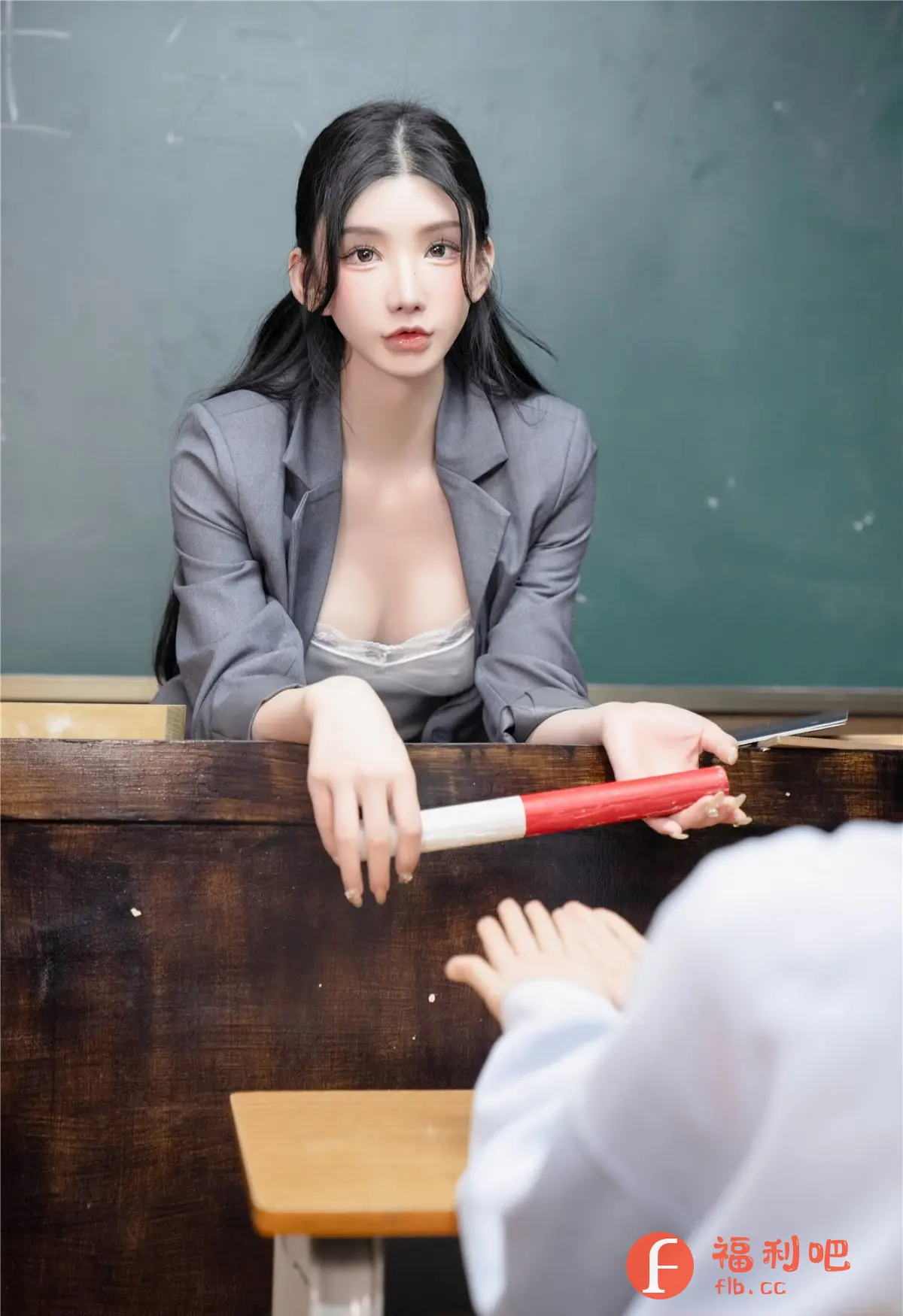这张照片展现了一位年轻女性,她身穿一套灰色的西装外套,内搭一件带有蕾丝边的白色内衣,略显性感。她留着一头乌黑的长发,发型精致,衬托出她精致的五官和白皙的皮肤。她的妆容非常精致,眼睛大而有神,嘴唇红润饱满。她站在一张老旧的木质课桌前,背景是绿色的黑板,营造出一种教室的氛围。她的手扶着桌沿,手中拿着一个红白相间的教鞭,似乎正在进行教学。课桌的另一边可以看到学生的手,仿佛正在接受指导。整个画面构图巧妙,光线柔和,凸显了女性的美丽和知性,也带有一丝性感和挑逗。照片整体风格偏向私房写真,可能旨在展现女性的魅力和自信。