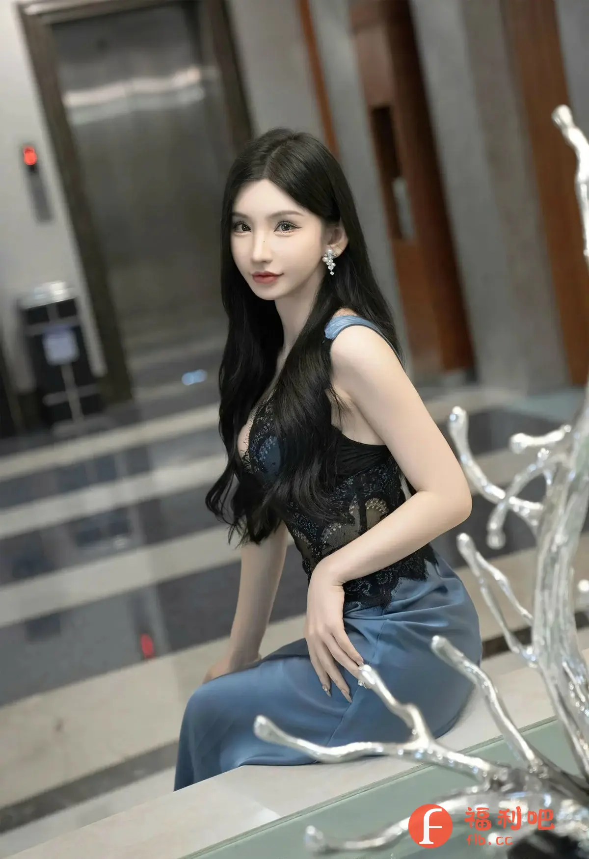 描述：照片中是一位年轻的亚洲女性，她有着精致的妆容，一头乌黑亮丽的长发自然地披在肩上。她身穿一件带有黑色蕾丝装饰的蓝色丝绸质感的连衣裙，裙子的设计凸显了她的身材曲线。她的左手轻放在裙子上，手指修长，指甲上装饰着精致的指甲彩绘。她佩戴着一对闪亮的耳环，增添了几分优雅气质。

背景较为模糊，可以看到电梯以及条纹装饰，暗示着她可能身处一个高档场所。画面右侧的银色树枝状装饰物增添了艺术感和视觉趣味。整体而言，这张照片展现了一种成熟女性的魅力和时尚品味。