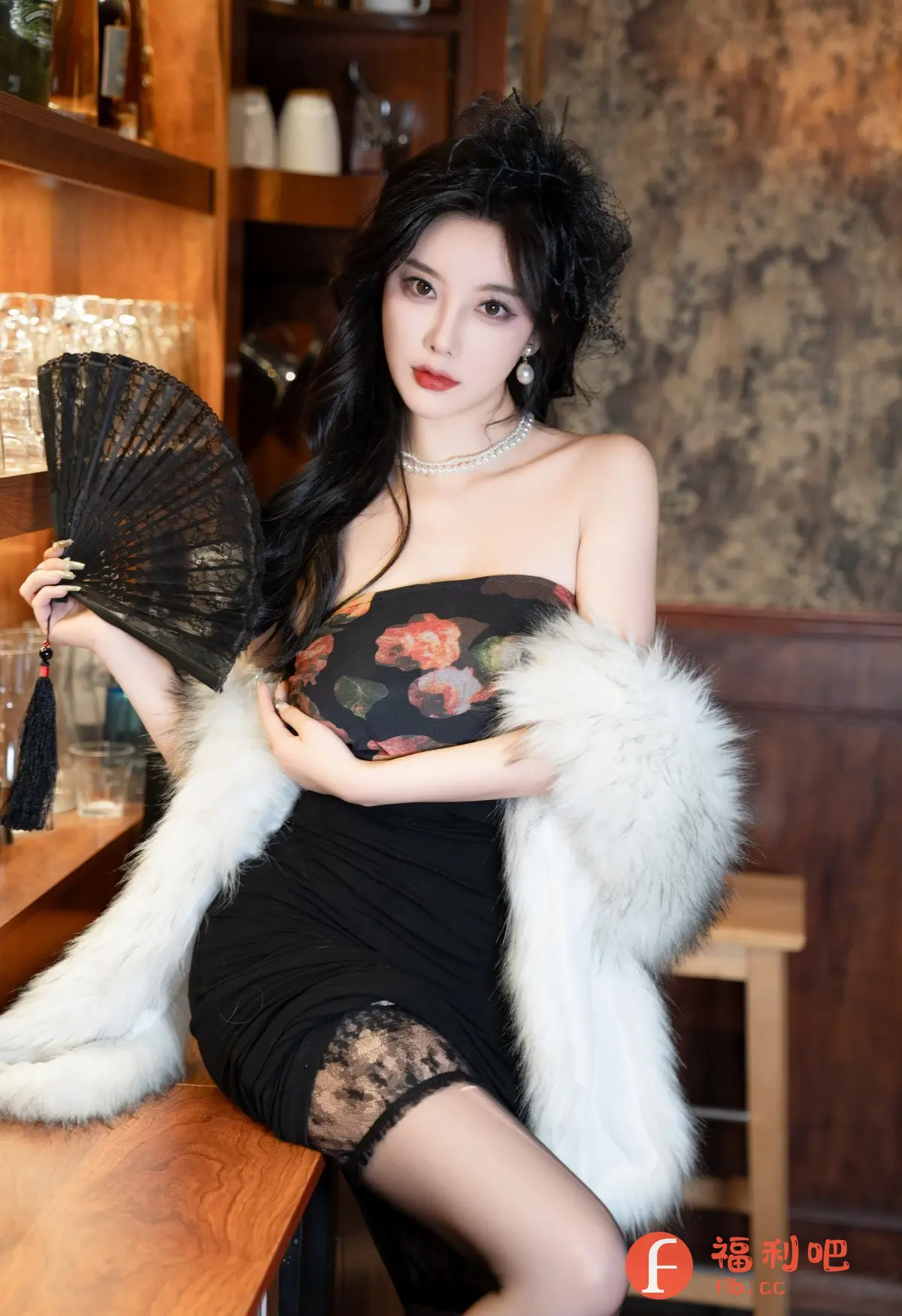 这张照片是一位穿着时髦的年轻女性的私房写真。她穿着一件黑色紧身连衣裙，上面搭配了一件无肩带的印花上衣，以及一件白色毛茸茸的披肩，显得既性感又优雅。连衣裙的下摆处露出了一点蕾丝花边，增添了一丝妩媚。她戴着珍珠项链和耳环，头上还装饰着一个黑色的发饰。她手持一把黑色的扇子，姿态优雅。背景是一个装饰着酒瓶和玻璃杯的吧台，以及带有复古花纹的墙壁，营造出一种奢华的氛围。整体来看，这张照片充满了时尚感和诱惑力。