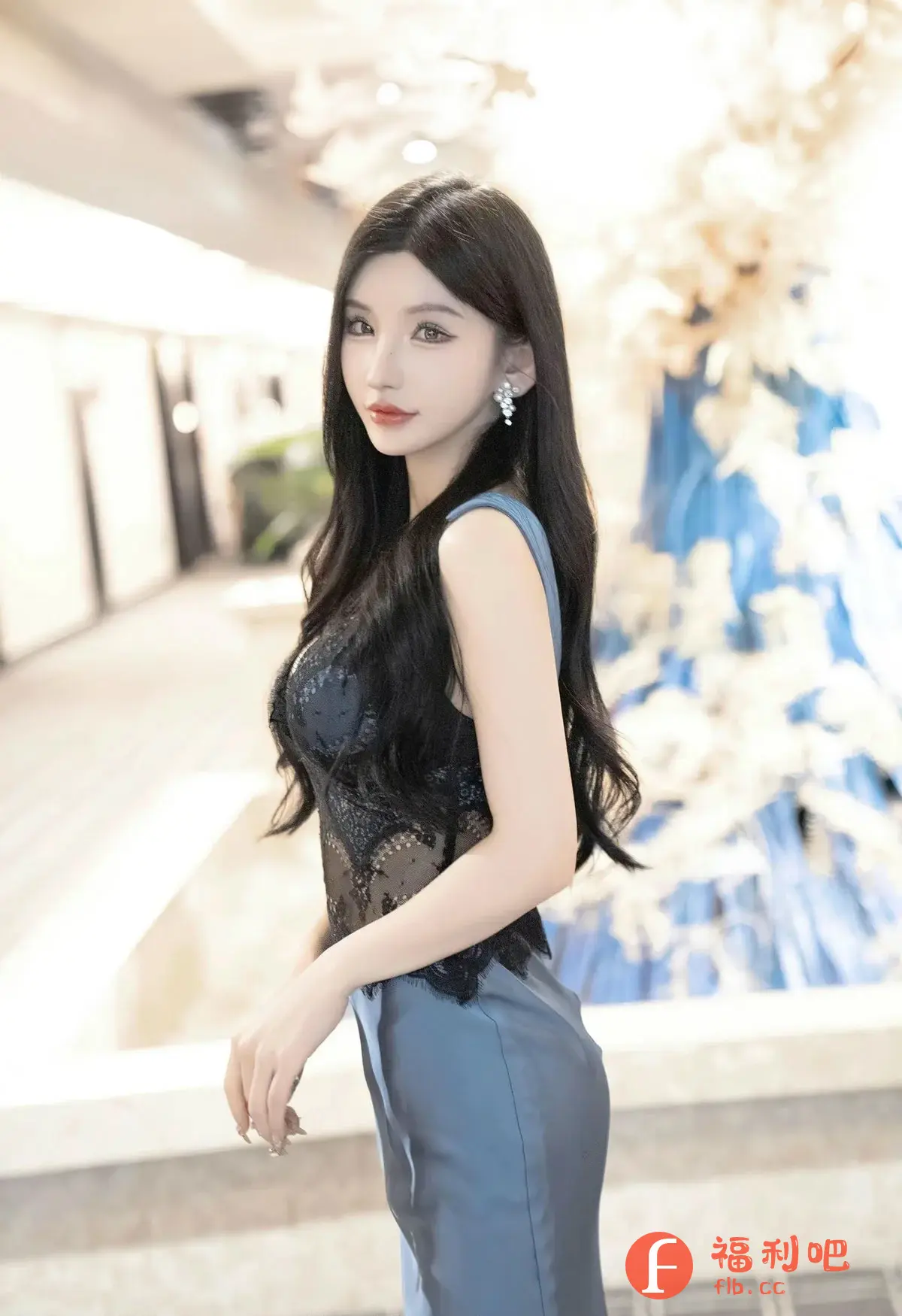 描述：这位年轻的亚洲女性身穿一件精致的黑色蕾丝上衣，内衬隐约可见，搭配一条光滑的蓝色丝绸裙子，优雅地站在室内环境中。她拥有一头乌黑亮丽的长发，自然地披在肩上，衬托出她精致的五官和白皙的皮肤。她的妆容淡雅，凸显了她的自然美，唇色饱满，更增添了一丝妩媚。她佩戴着闪耀的耳环，为整体造型增添了一份奢华感。她微微侧身，眼神温柔地注视着镜头，展现出自信和迷人的魅力。背景模糊，但可以辨认出是酒店或豪华场所的走廊，装饰着精美的花艺，营造出一种高雅的氛围。整体而言，这是一张充满时尚感和女性魅力的照片。