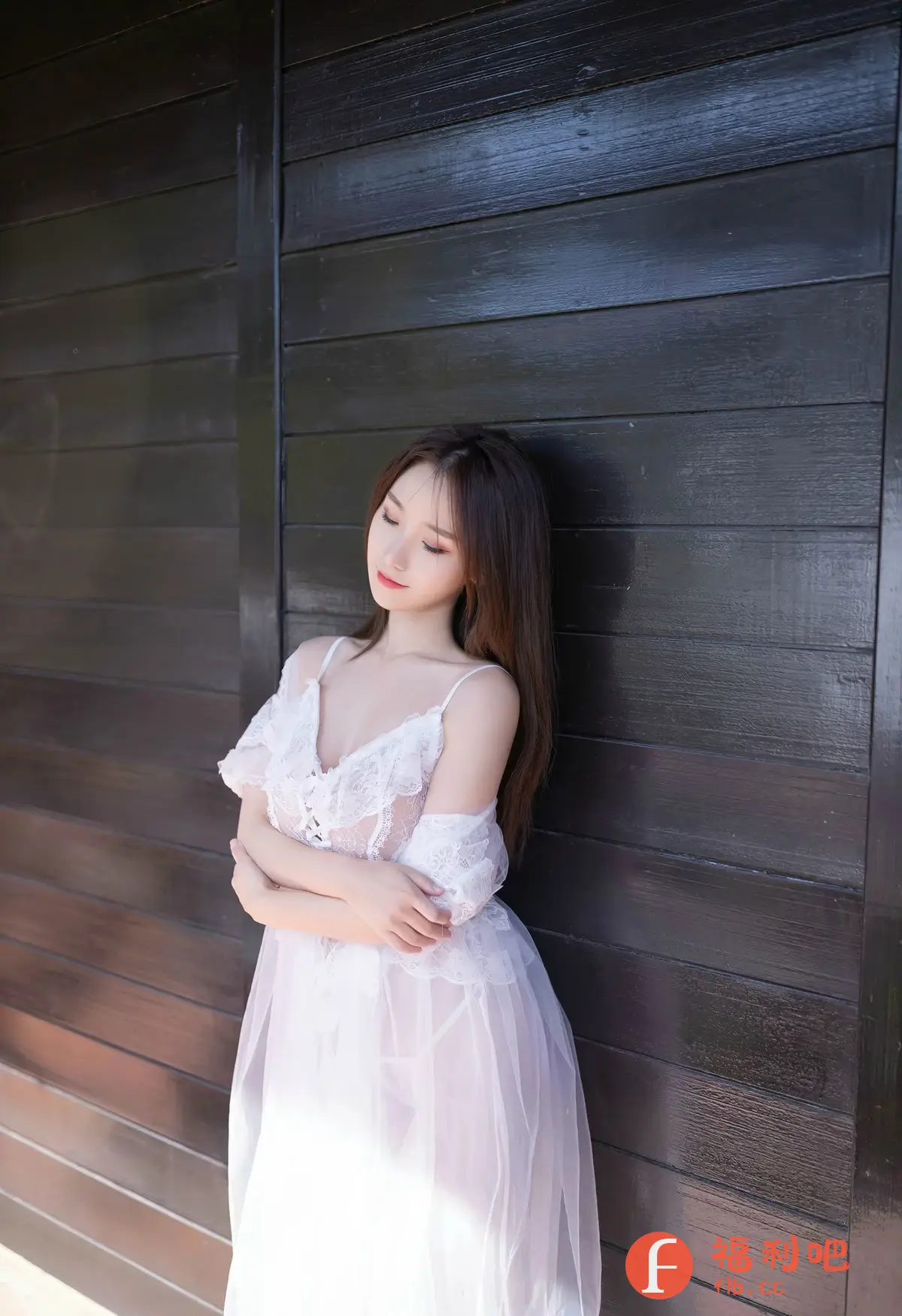 照片中是一位年轻美丽的亚洲女性，她身穿一件白色的蕾丝薄纱长裙，裙子的设计带有吊带和低胸，透露出一种性感和柔美的气息。她微微闭着眼睛，神情恬静，仿佛沉浸在自己的世界里。她的双手交叉放在胸前，姿态优雅而放松。她的长发自然地垂落在肩上，发质柔顺，颜色乌黑，与白色的裙子形成鲜明的对比。她背靠着一堵深色的木质墙壁，墙壁的纹理清晰可见，为照片增添了一种复古和质朴的氛围。整体光线柔和，营造出一种温馨和浪漫的氛围，突显了女性的柔美和性感。这是一张充满艺术感的私房写真，展现了女性的魅力和内在的平静。