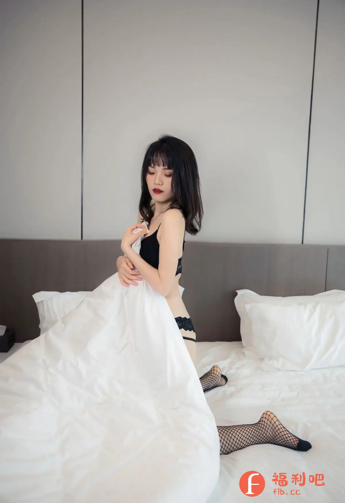 照片中,一位亚裔女性跪坐在床沿上,她穿着一件黑色吊带背心和带有蕾丝边的黑色内裤,身上盖着洁白的被子,露出一双穿着黑色鱼网袜的脚。她的头发是黑色短发,刘海垂在眉毛上方,红色的嘴唇显得格外醒目。她的眼神向下,表情略带忧郁,似乎在思考着什么。背景是浅灰色的墙壁和棕色的床头板,营造出一种简约而私密的氛围。整个画面充满了诱惑力,同时也展现了女性的柔美和性感。