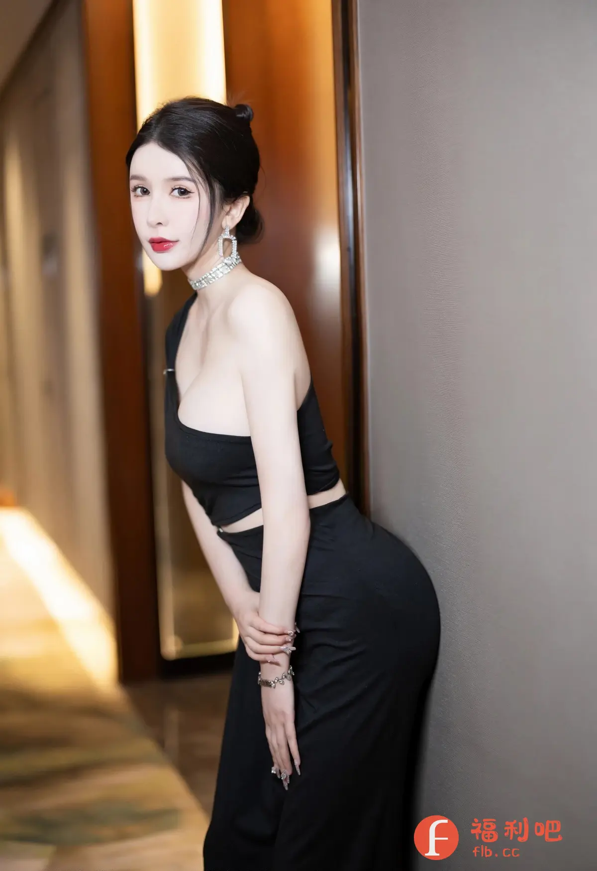照片中一位年轻女性倚靠在墙边,穿着一件时尚的黑色露肩套装,上身是短款的单肩上衣,露出部分腰部,下身则是高腰的直筒裙,勾勒出她的曲线。她佩戴着闪亮的项链和手链,耳朵上戴着大而华丽的耳环,增添了几分奢华感。她的头发盘起,露出精致的脸庞和红唇,眼神迷人,姿态优雅。整体风格简约而性感,展现出女性的魅力和自信。