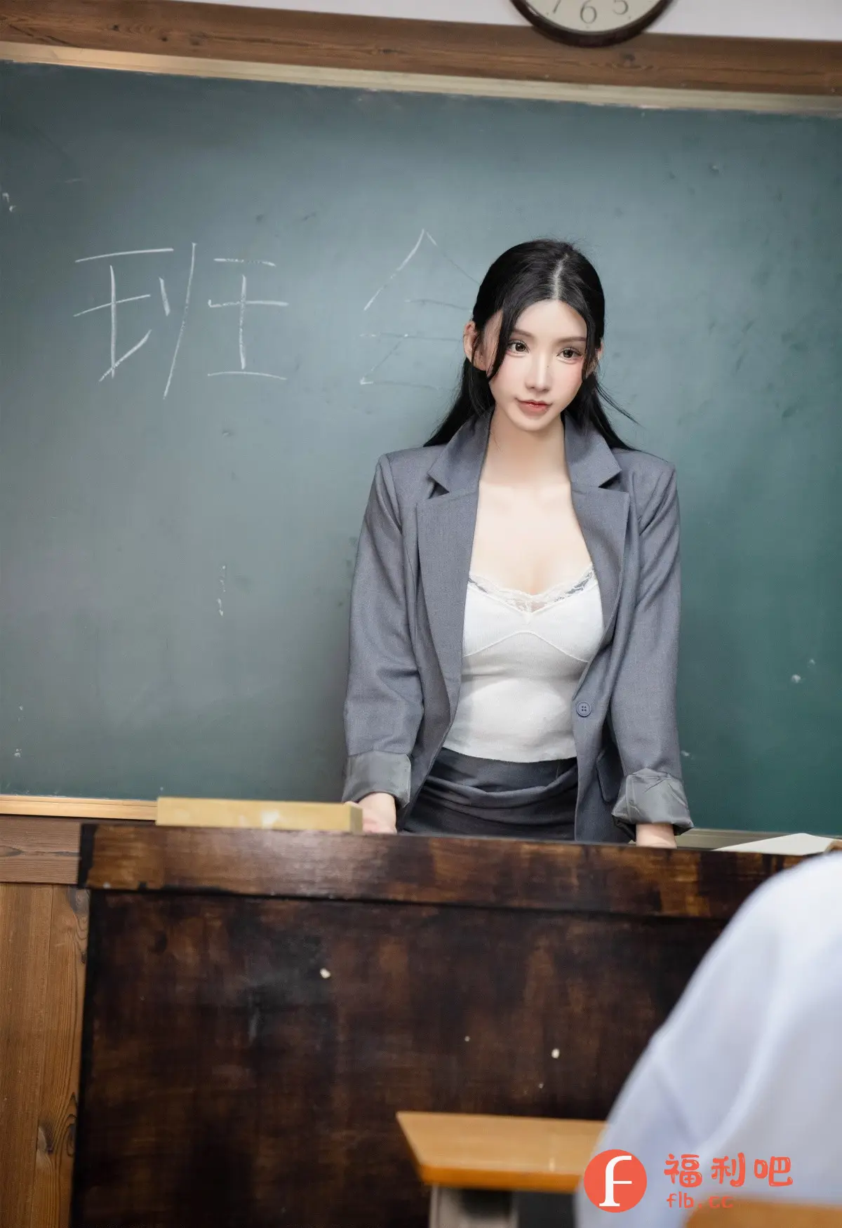 这张照片是一位年轻女性的个人写真,她站在教室的黑板前,穿着一件灰色的西装外套,内搭一件白色蕾丝边的背心,下身穿着一件深色的裙子,整体着装显得既专业又略带性感。她的长发自然地披散在肩上,皮肤白皙,妆容精致,眼神略带忧郁,给人一种知性成熟的感觉。黑板上用白色的粉笔写着一些文字,可能是课堂内容或者班级事务。画面整体光线柔和,营造出一种静谧的氛围,可能是在课后或者课间休息的时候拍摄的。从服装和场景来看,这位女性很可能是一位老师或者教育工作者,拍摄场景很可能是一家教室或者教育机构。