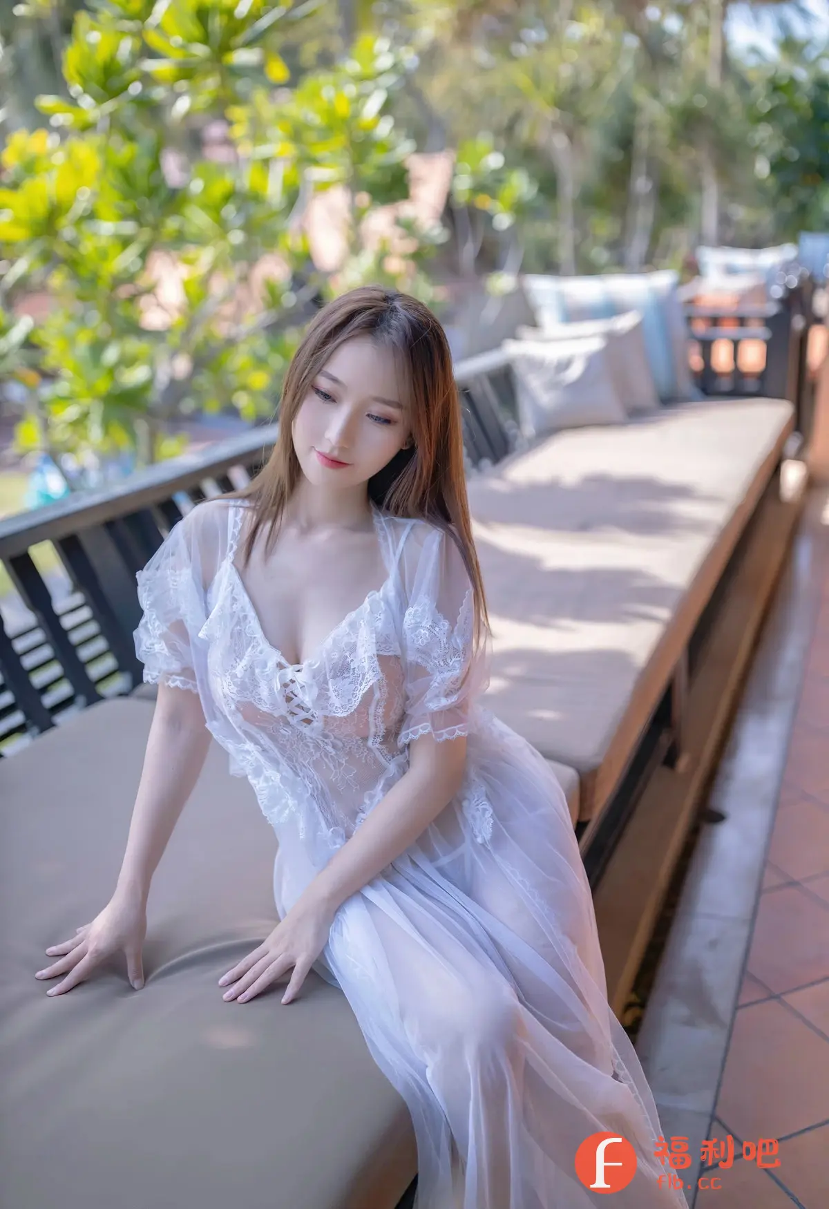 这张照片展现了一位年轻女性的私房写真，她身穿一件轻薄的白色蕾丝长裙，裙子透明的设计隐约显露出她的肌肤，增添了一丝性感和神秘感。她的长发柔顺地披在肩上，微微低头，眼神温柔，流露出一种慵懒和放松的氛围。她坐在一个长长的沙发或躺椅上，背景是户外的景色，有绿色的植物和阳光，营造出一种轻松舒适的度假氛围。整体构图和光线运用都非常专业，突出了女性的美丽和优雅。
