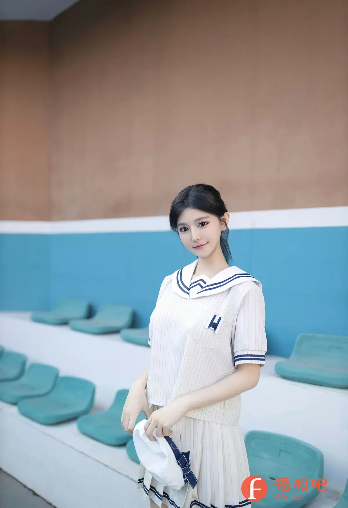 描述:照片中,一位年轻女性穿着一件白色的水手服套装,套装的细节包括蓝色的条纹和领口,以及裙子上的蓝色蝴蝶结。她的头发乌黑亮丽,扎成一个简单的发髻,露出了精致的五官。她手持一顶白色的帽子,帽子上也有蓝色的装饰,与她的服装相呼应。背景是一个带有蓝色座椅的看台,上方是棕色的墙面。整体光线柔和,营造出一种清新自然的氛围。她的表情温婉,带着一丝微笑,给人一种亲切可人的感觉。整个画面充满了青春活力,展现了女性的柔美和自信。