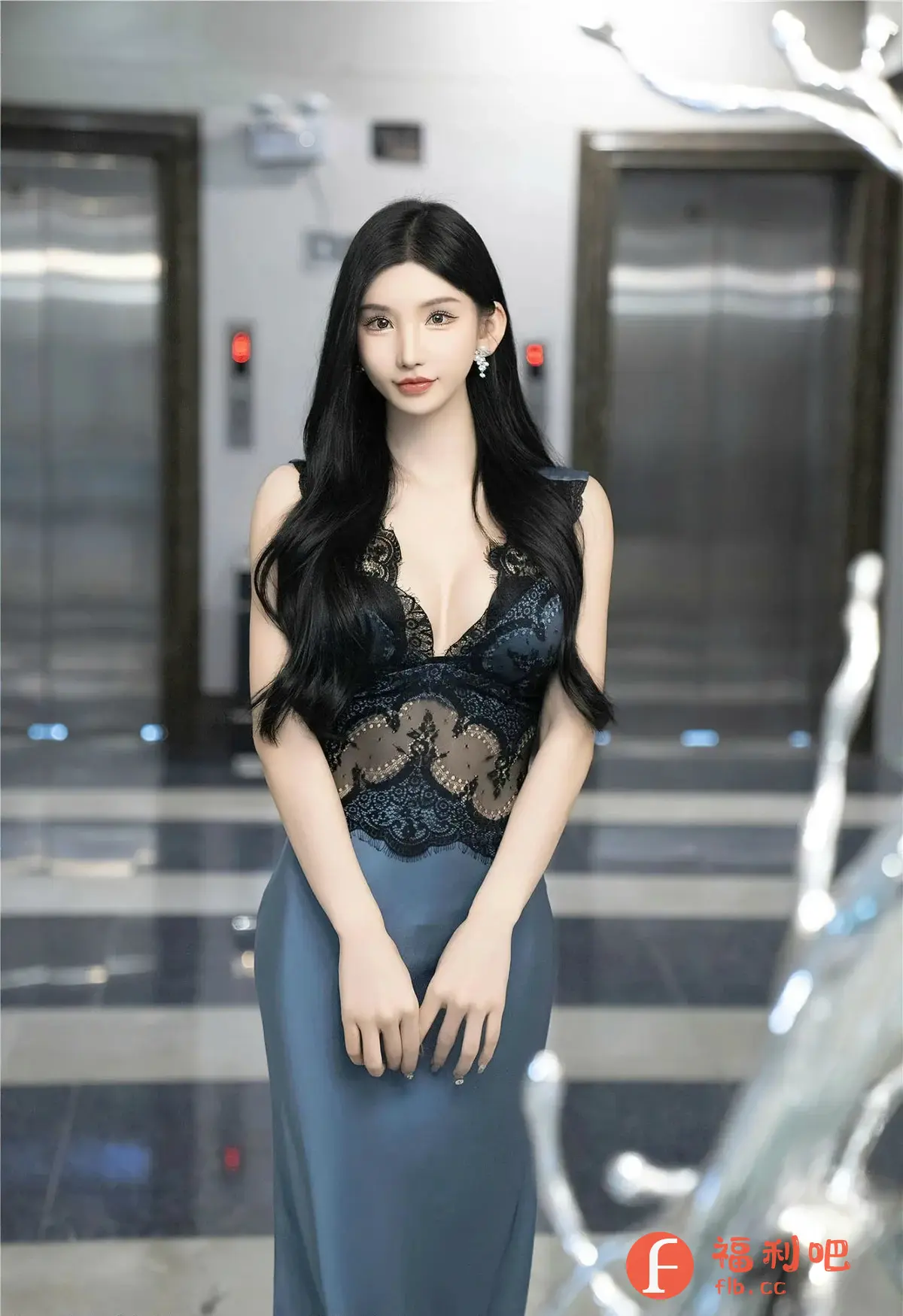 这张照片是一位年轻女性的私房写真，她站在一个看起来像是酒店大堂的场所，背景是闪亮的电梯和具有现代感的设计。她身穿一件深蓝色吊带裙，上身是精致的黑色蕾丝，增加了性感的元素，同时也衬托出她白皙的肤色。她的长发乌黑亮丽，自然地垂在肩上，一侧还别着闪耀的耳饰，为整体造型增添了亮点。她的双手轻放在身前，姿态优雅而自信，眼神平静而迷人。整体的光线柔和，营造出一种温馨而私密的气氛，凸显了女性的柔美气质，体现了拍摄者捕捉美的独特视角。