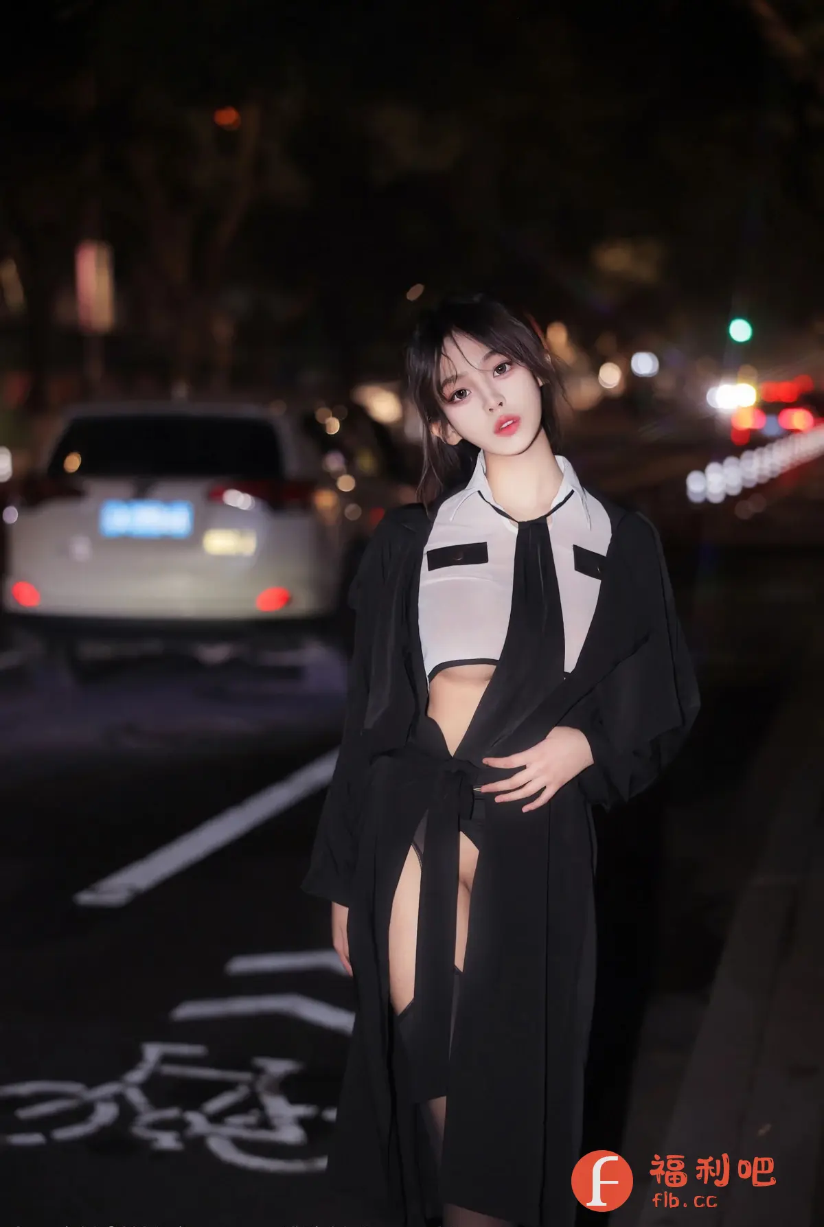 照片中是一位年轻女性,她站在夜晚的街头,背景中可以看到模糊的车辆和灯光。她穿着一件黑色长款外套,内搭一件白色短款上衣,露出腹部。外套敞开,腰间系着腰带,下身搭配黑色短裤,腿部穿着黑色丝袜。她的头发扎起,脸部妆容精致,眼神看向镜头,姿态优雅而自信。整体风格性感而时尚,凸显了女性的魅力和个性。