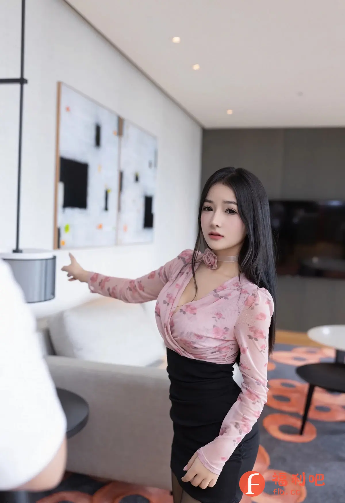 这位美女身穿一件别致的服装，上半部分是粉色花纹的薄纱上衣，设计性感而优雅，下半部分则是黑色紧身裙，凸显了她的身材曲线。她的长发乌黑亮丽，自然地垂落在肩上，增添了几分妩媚。美女佩戴了一条与上衣颜色相呼应的颈链，更显精致。她站在室内，背景是现代简约风格的装修，墙上挂着抽象画作，衬托出她的时尚品味。美女似乎正对着镜头微笑，眼神温柔，给人一种亲切的感觉。整体画面充满了生活气息，展现了美女自信、美丽的一面。
