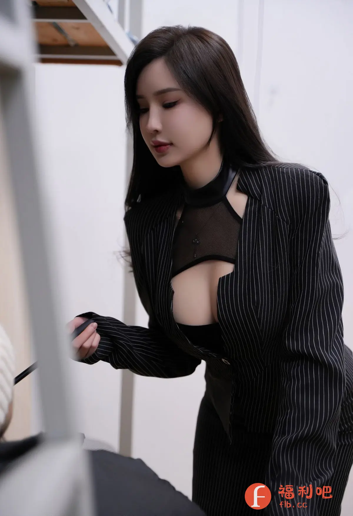 照片中,一位身材姣好的年轻女性身穿一件设计大胆的黑色条纹西装,内搭一件带有黑色皮质领口的镂空网纱上衣,露出迷人的事业线。她一头乌黑亮丽的长发自然垂落在肩头,精致的妆容更显妩媚动人。她的眼神略带忧郁,仿佛在思考着什么。她手持一个黑色物体,似乎正在专注于某项工作。照片的背景简洁,以白色为主,更衬托出她的美丽和优雅。整体画面充满时尚感和神秘感,令人不禁想要了解更多关于她的故事。