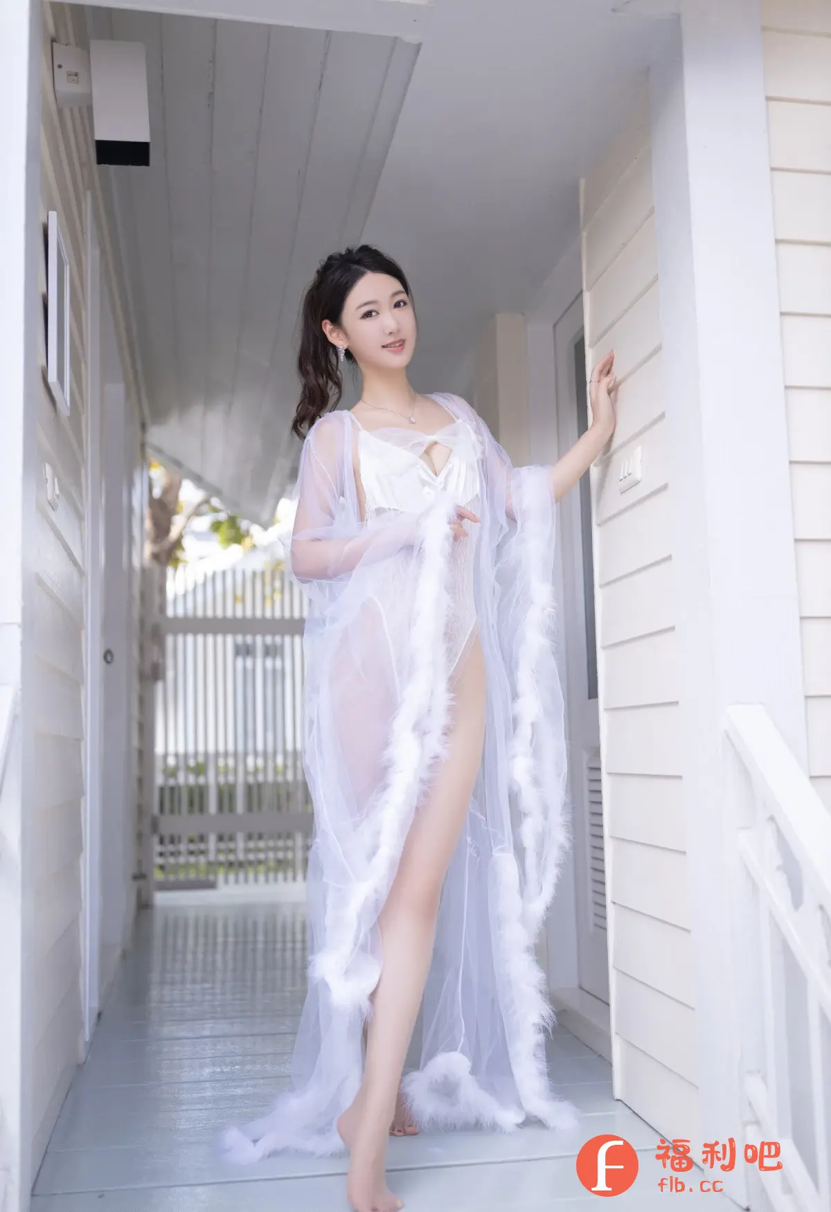照片中是一位穿着白色内衣和透明长袍的亚洲女性。她长发扎成马尾,戴着耳环和项链。她站在一个白色的走廊里,一只手扶着墙,姿势优雅。长袍边缘有羽毛装饰,增添了轻盈感。走廊尽头可以看到室外栅栏和树木,背景简洁而明亮,突出了女性的柔美和性感。