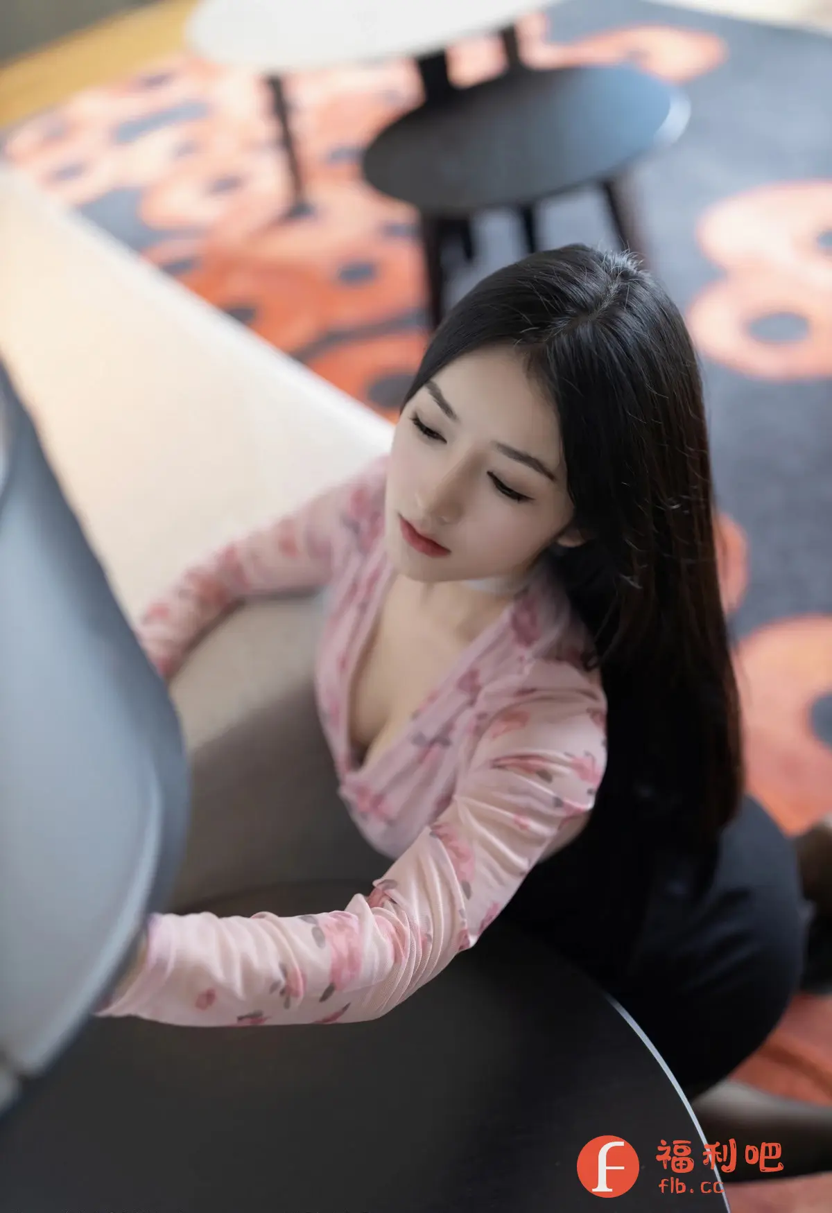 一位身穿粉色印花上衣的美女出现在镜头前，她的长发自然垂落，增添了几分妩媚。她微微低头，眼神温柔而专注，仿佛沉浸在自己的世界里。上衣的V领设计，若隐若现地展现了她的锁骨线条，更显性感。背景中，柔软的沙发和别致的茶几营造出一种舒适而温馨的氛围。整体画面充满了女性的柔美与优雅，让人感受到一种宁静和放松。