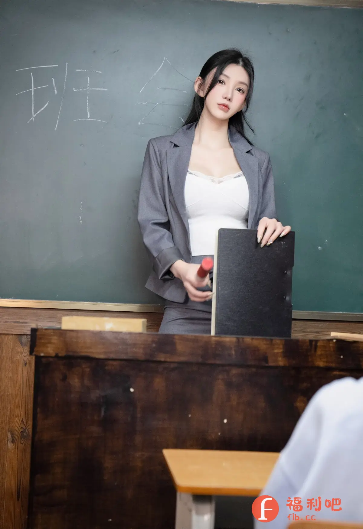 描述:一位年轻女性站在黑板前,身穿灰色西装外套和白色内搭,下身搭配同色系裙子,展现出一种既专业又时尚的风格。她的长发自然垂落,妆容精致,眼神专注,手中拿着教案和教鞭,似乎正在准备上课或进行讲解。黑板上写着一些汉字,可能是课程内容或班级名称。场景设置在一个教室里,课桌椅和木质墙面营造出一种传统的学习氛围。整体画面展现出一种知性美和职业感。