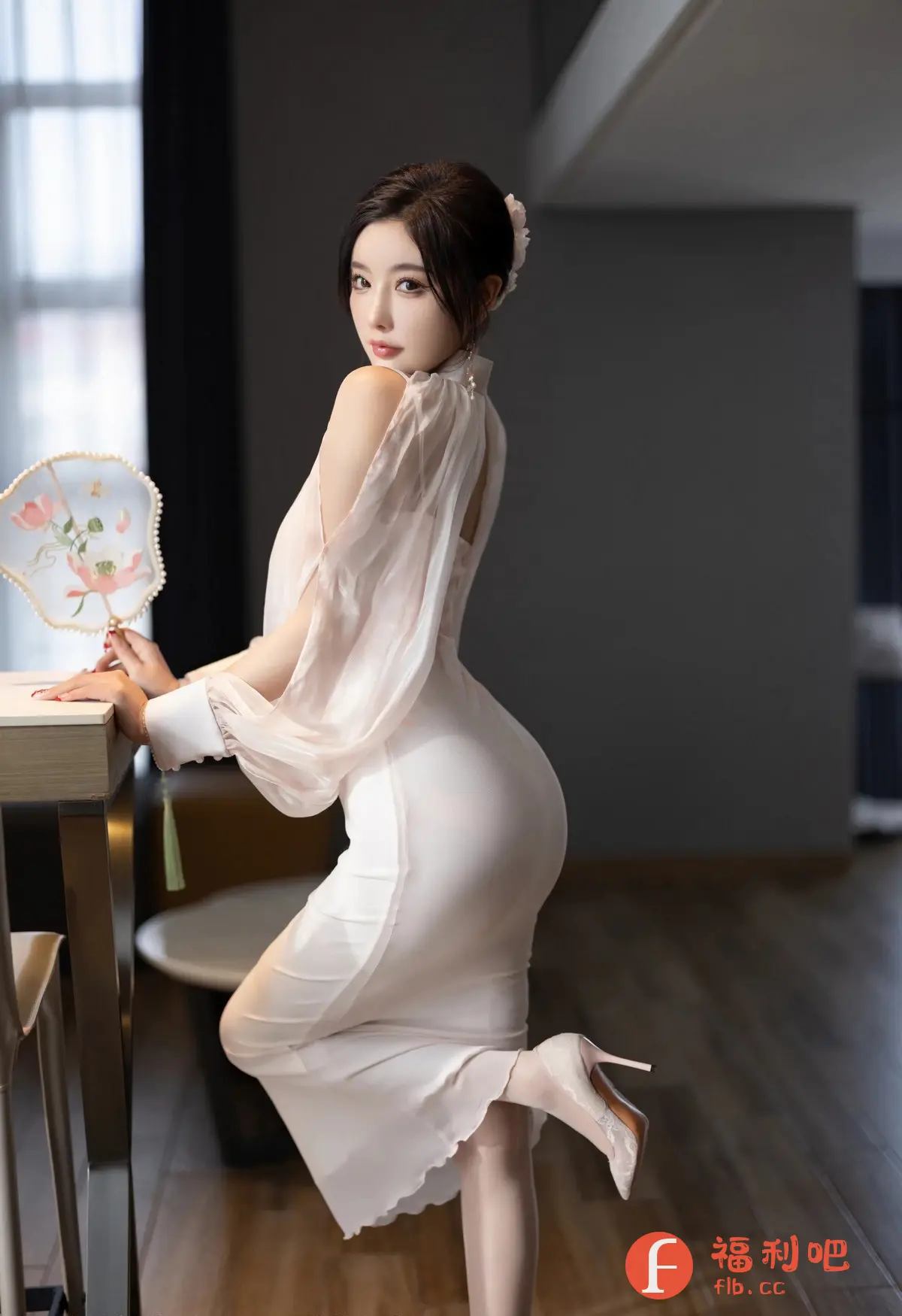 照片中，一位年轻貌美的亚洲女性身穿一袭优雅的白色连衣裙，裙子的设计别致，上半身搭配了轻盈的薄纱长袖，更显仙气飘飘。她侧身站在桌旁，一手扶着桌面，另一手轻握一把精致的团扇，扇面上绘有淡雅的莲花图案。她的发型古典精致，盘发上点缀着一朵白色的小花，更增添了她的柔美气质。她回眸一笑，眼神清澈动人，仿佛诉说着一个美丽的故事。她脚上穿着一双浅色的高跟鞋，更加凸显了她修长的身材。背景是室内环境，光线柔和，营造出一种温馨浪漫的氛围。整体画面唯美典雅，展现了女性的柔美和高贵。