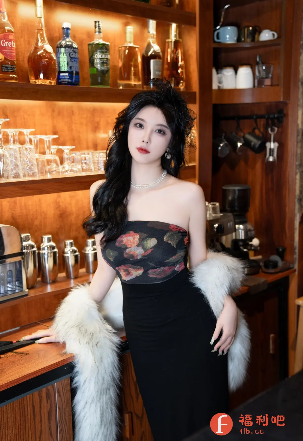 描述：这位美女身着一袭紧身黑色礼服，上身是带有花朵图案的抹胸设计，尽显优雅气质。她佩戴着珍珠项链和精致的耳环，头上还装饰着一朵小小的黑色羽毛。美女披着一件白色的毛绒披肩，更添一份奢华感。她站在一个装饰着各种酒瓶、杯具和咖啡机的背景前，似乎是在一个高级酒吧或咖啡馆里。她的妆容精致，红唇鲜艳，眼神迷人，散发着一种成熟女性的魅力。整体照片的光线柔和，突出了美女的精致五官和优美的身姿，营造出一种私密而又浪漫的氛围。
