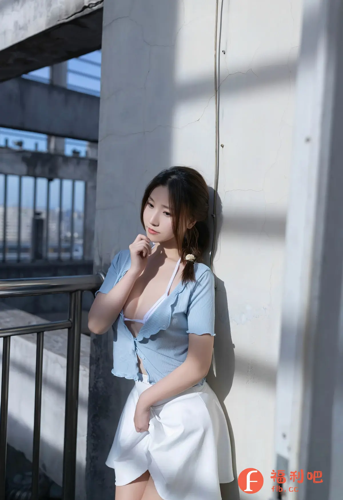 一位年轻女性站在户外的栏杆和墙边,她穿着一件敞开的浅蓝色短款上衣,内搭白色比基尼,下身是一条白色短裙。她微微侧着身子,一只手放在下巴附近,似乎在思考什么,视线则看向左侧。她的头发扎成低马尾,搭配一个简单的发饰。阳光洒落,在墙面上形成了明暗对比,为画面增添了层次感。背景中可以看到建筑结构和城市景观,暗示了照片拍摄于较高的位置。整体风格清新自然,展现了女性的青春活力和性感魅力。