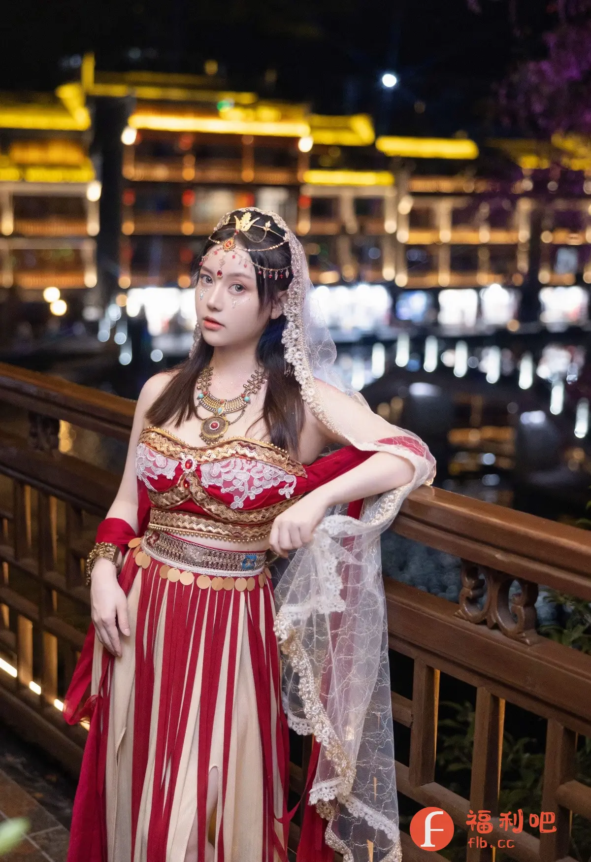 照片中,一位美丽的年轻女性身着华丽的传统服饰,站在夜晚的户外。她有一头棕色的长发,戴着精致的头饰,上面镶嵌着宝石,面部也化着精致的妆容,眼睛下方点缀着亮片。她身穿一件红白相间的长裙,上身是刺绣精美的红色束胸,腰间系着装饰着亮片的腰带。她的脖子上戴着一条华丽的项链,手臂上戴着手镯。她倚靠在木制的栏杆上,一只手轻轻地扶着一条白色的纱巾,纱巾上装饰着金色的蕾丝。背景是灯火通明的建筑群,灯光在水面上反射,营造出一种浪漫而神秘的氛围。整体画面色彩鲜艳,细节丰富,展现了女性的美丽和服饰的华丽。