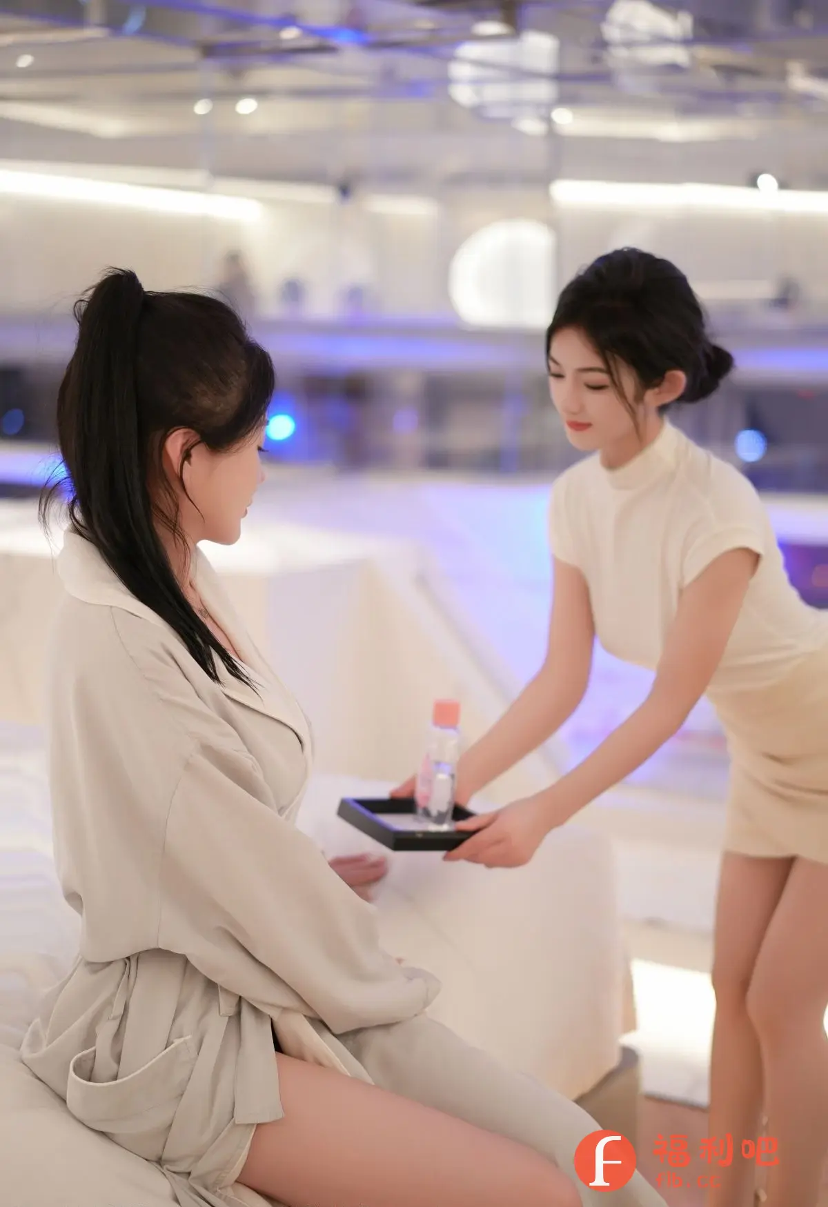 这张照片展现了两位年轻女性的私房写真场景。一位女性身穿宽松的米色睡袍,优雅地坐在床上,长发扎成高马尾,露出精致的侧颜,眼神温柔,似乎在欣赏着什么。另一位女性则穿着浅色套装,短裙展现了她修长的双腿,她正端着一个黑色托盘,上面放着一瓶矿泉水,准备递给床上的那位女性。整个画面色彩柔和,灯光温暖,营造出一种舒适、放松的氛围。背景是模糊的室内环境,可能是酒店或高级公寓的卧室,整体给人一种高品质、精致的生活体验。照片展现了女性的柔美和优雅,同时透露出一种私密、亲近的氛围。