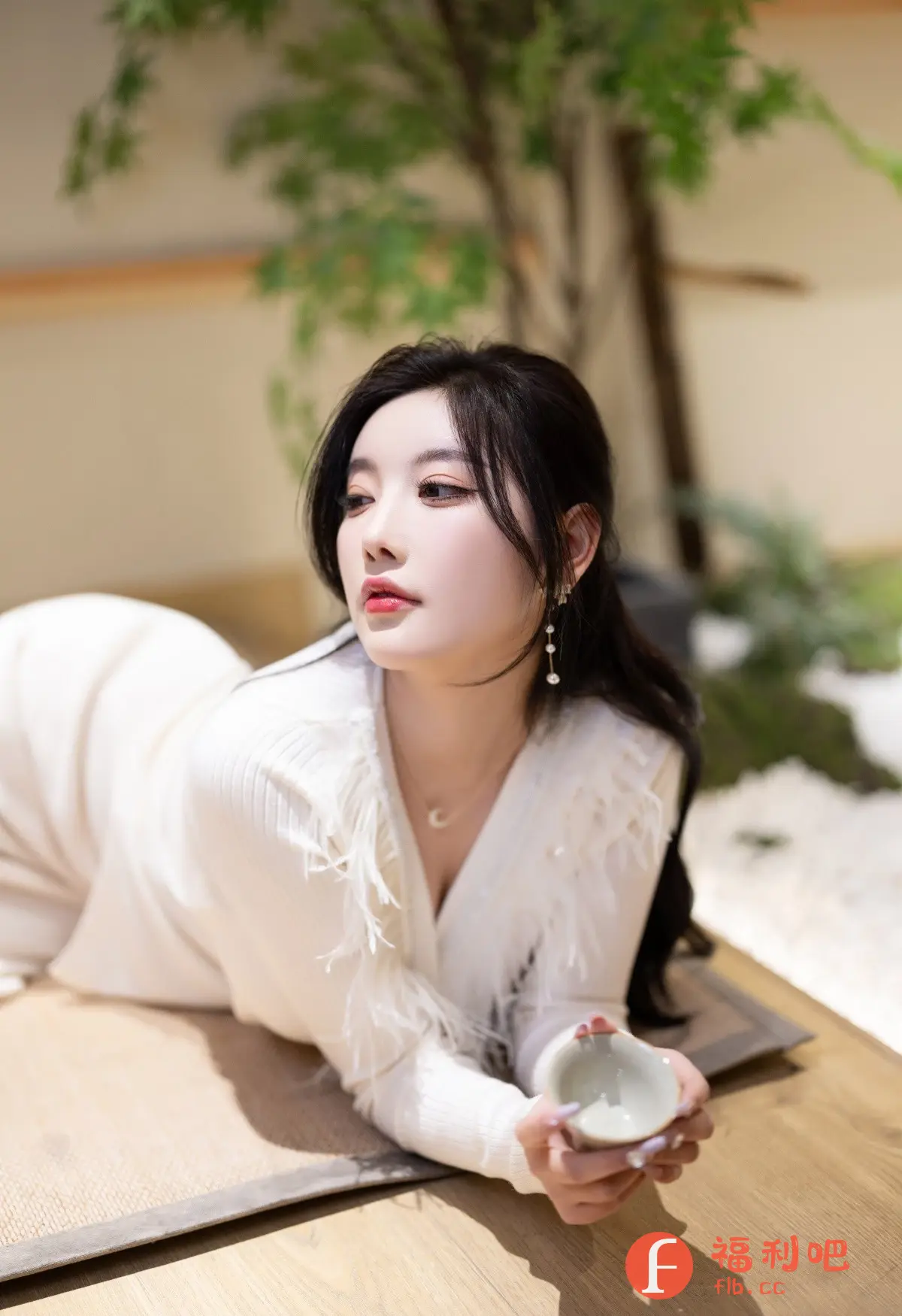 这是一张美女的私房写真照片,背景是雅致的室内环境,可以看到一些绿植,营造出一种清新自然的感觉。美女穿着一件白色的针织连衣裙,裙子上点缀着羽毛,显得温柔又时尚。她侧身躺在铺着席子的木质地板上,姿态慵懒而优雅。她的头发乌黑柔顺,佩戴着精致的耳饰,增添了几分精致感。她手捧着一个小茶杯,眼神温柔,表情恬静,给人一种岁月静好的感觉。整体画面清新唯美,展现了美女的优雅气质和温柔魅力。