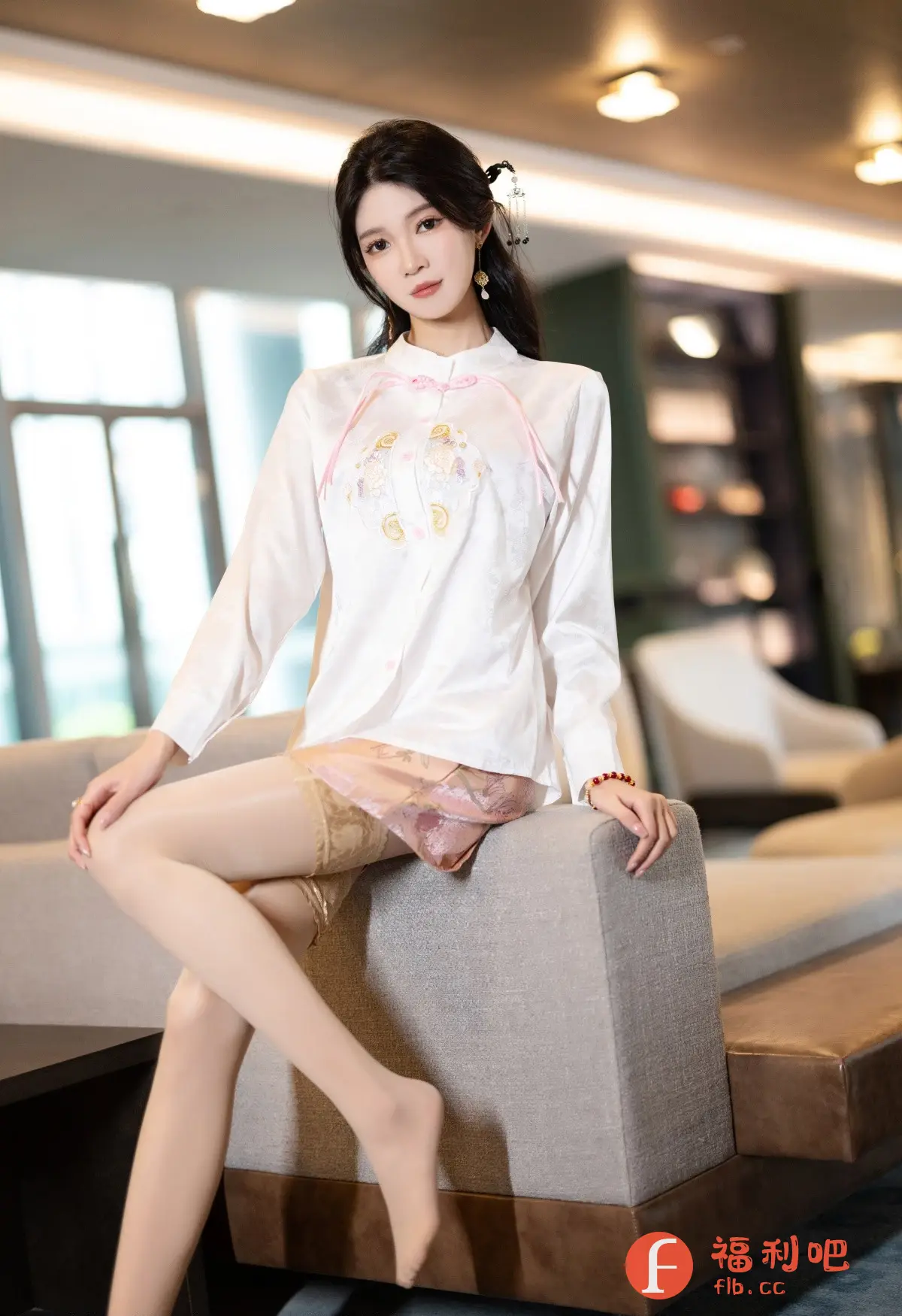 这张照片是一位美女的私房写真,她坐在沙发上,姿态优雅。她穿着一件丝质白色长袖上衣,上衣领口有粉色的盘扣和绣花图案,搭配一件带有蕾丝花边的粉色短裙,更显妩媚动人。美女一双修长的腿包裹着肉色丝袜,光脚踩在地毯上,增添了一丝慵懒的气息。她的头发盘起,发髻上别着精致的发饰,耳朵上带着流苏耳环,更衬托出她精致的五官。照片背景是现代风格的房间,透过窗户可以看到室外的景色,整体营造出一种时尚而又充满东方韵味的气氛。无论是她的穿着、妆容,还是姿态,都展现出一种独特的东方女性魅力。