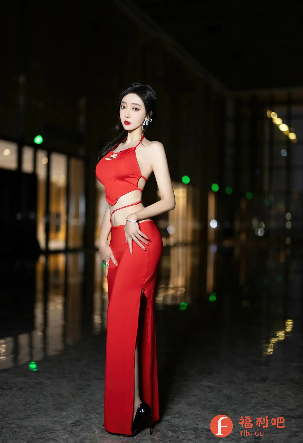这张照片展示了一位年轻女性，她身着一套引人注目的红色两件套服装。这套服装包括一件露脐上衣和一条开衩的长裙，凸显了她的身材。她穿着一双黑色的高跟鞋，增加了整体造型的优雅感。她的头发整齐地向后梳，露出精致的五官，并佩戴着一对闪闪发光的耳环。她的妆容很精致，红色的嘴唇和深邃的眼神为她增添了魅力。照片的背景是室内环境，有一些模糊的光点，营造出一种时尚而神秘的氛围。整体的构图和光线都突出了模特的个人魅力和服装的时尚感。