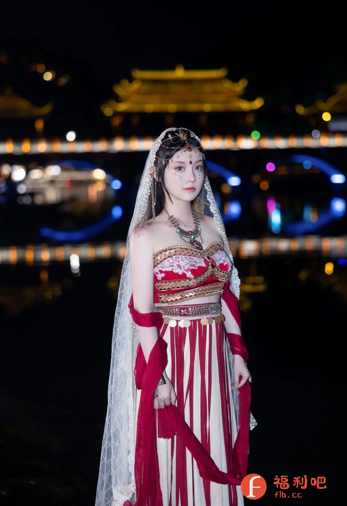 照片中,一位美丽的亚洲女性身着华丽的传统服饰,站在一个背景模糊的夜晚。她的服饰颜色鲜艳,以红色、白色和金色为主,包括一个精致的头饰和面纱。她的妆容也很精致,脸上贴着闪亮的装饰品。背景是一个带有灯光的桥梁和建筑物,水面反射出灯光,营造出一种梦幻般的气氛。整体来看,这是一张精心拍摄的私房写真,突显了女性的美丽和服饰的华丽,同时也利用背景营造出一种神秘和浪漫的氛围。