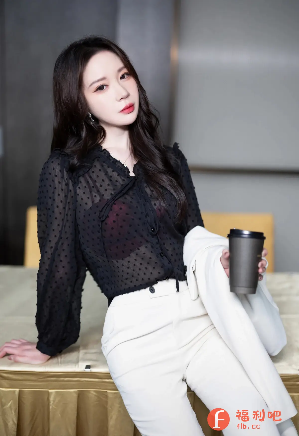 照片中是一位年轻女性,她穿着时尚且略带性感。她身穿一件黑色的透明波点衬衫,内搭一件红色内衣,下身搭配一条白色的紧身长裤,勾勒出她的身材曲线。她手中拿着一个黑色咖啡杯,另一只手拿着一件白色外套,随意地搭在腿上。她的头发乌黑亮丽,自然地披散在肩上,精致的妆容凸显出她美丽的容貌。她的眼神略带慵懒,给人一种成熟妩媚的感觉。整体造型优雅而时尚,展现了她自信和魅力。背景看起来像是在室内,可能是酒店或者办公室,带有现代简约的风格。