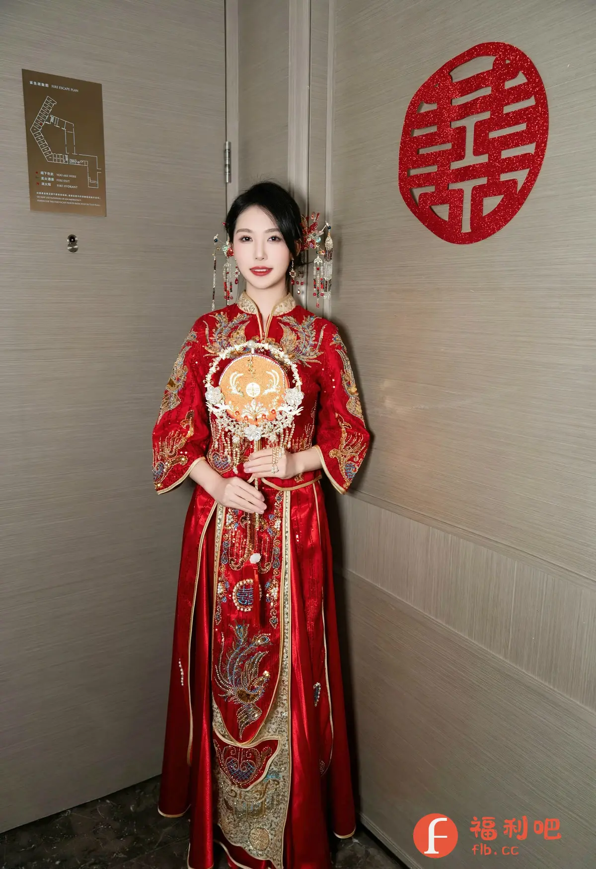 照片中,一位东方女性身着华丽的中式红色礼服,手持精致的团扇,仪态端庄地站立在室内。礼服上绣有精美的传统图案,色彩鲜艳,细节丰富,彰显了传统中式婚礼的喜庆氛围。她的头发盘起,佩戴着古典的发饰,更增添了古典韵味。背景是现代风格的墙面,墙上贴着一个红色的“囍”字,寓意着喜庆和美满。整体画面充满了浓郁的东方风情,既有传统的美感,又不失现代的时尚感,展现了东方女性的优雅与魅力。