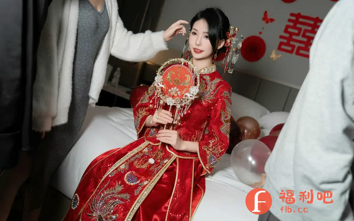 这张照片是一位穿着传统红色中式礼服的美丽女子。她坐在铺着白色床单的床上,手持一个精致的红色绣花扇子。她的头发盘起,并装饰着华丽的发饰,与她的礼服相得益彰。她的妆容精致,突显了她的美丽。背景中有一些红色的装饰品,墙上贴着红色的“囍”字,营造出喜庆的氛围。旁边有两个人,一个穿着灰色连衣裙和白色开衫,另一个穿着灰色衬衫。照片整体呈现出一种典雅、喜庆的感觉,很可能是在拍摄结婚相关的照片。