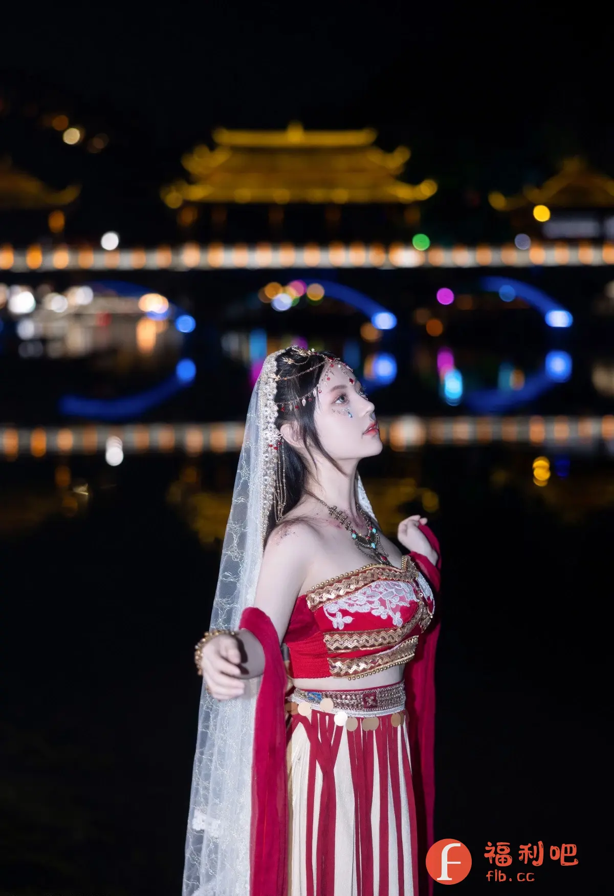一位年轻女子身着华丽的传统服饰,站在夜色下的水边。她抬头仰望,神情专注而充满期待。她的服装以红色和金色为主,搭配白色的面纱,更显其优雅气质。精致的头饰和颈饰增添了华丽感,脸上的妆容也十分特别,增添了她的神秘感。背景是灯光璀璨的建筑物和水面,倒映出迷人的色彩,为整个画面营造出梦幻般的氛围。整体而言,这是一张富有艺术感和文化气息的美丽照片。