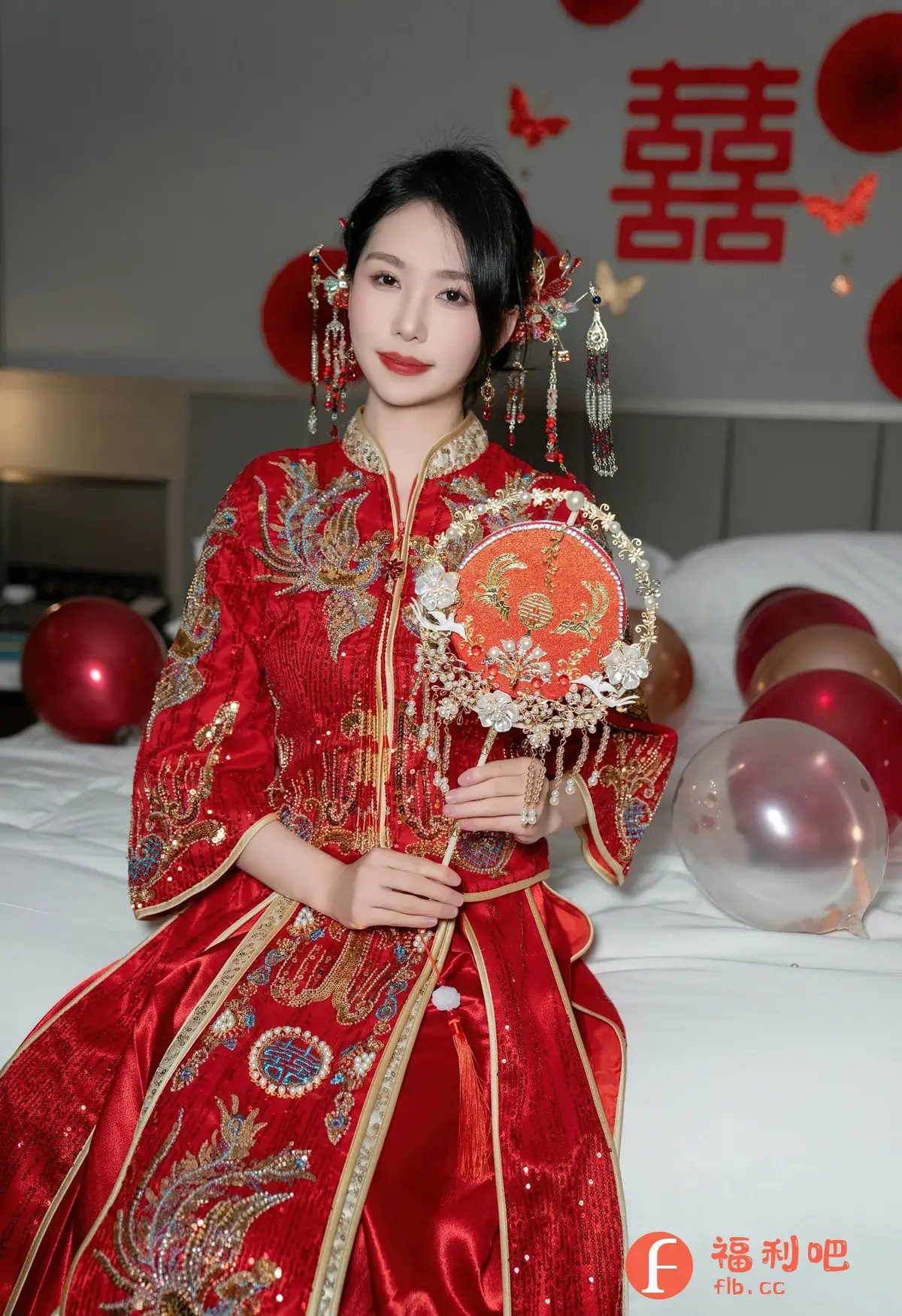 画面中是一位身着中式嫁衣的美丽女性。她坐在铺着白色床单的床上,周围装饰着红色和透明的气球,营造出喜庆的氛围。她的嫁衣是亮眼的红色,上面绣有精致的金色图案,显得华丽而隆重。她手中拿着一把装饰精美的团扇,扇面上也有喜庆的图案。她的头发盘起,并佩戴着传统的发饰,这些细节都体现了中式婚礼的传统元素。她的妆容精致,红唇更增添了一份妩媚。背景墙上贴有红色的喜字,进一步强调了婚礼的主题。整个画面充满了浓郁的中国风情,展现了新娘的美丽和幸福。