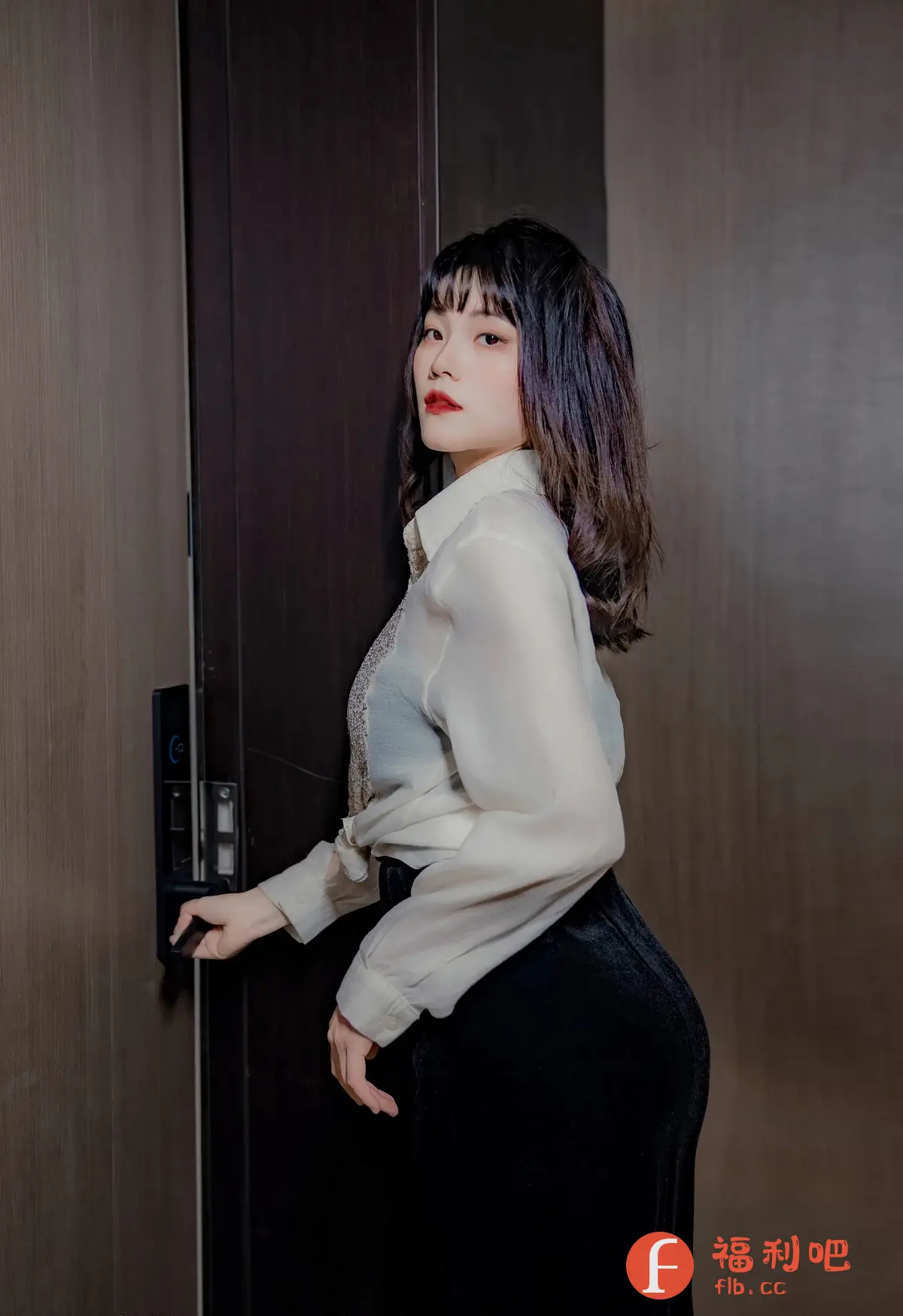这张照片是一位亚洲女性站在门口的私房写真。她有着乌黑亮丽的短发,齐刘海,精致的妆容,红唇十分醒目。她身穿一件半透明的白色衬衫,内衬一件带有装饰的内衣,下身搭配一条紧身黑色长裙,勾勒出优美的身材曲线。她的手放在门把手上,似乎正要推门进入。照片的背景是深色的木质墙面,营造出一种神秘而诱人的氛围。整体光线柔和,突出了女性的皮肤质感和身材曲线,展现了她的自信和魅力。