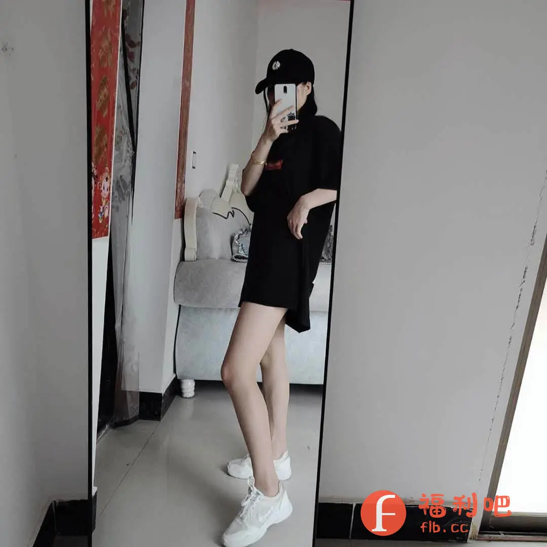 这位美女展示了一件黑色宽松T恤,看起来像连衣裙一样。她搭配了一顶黑色棒球帽和一双白色运动鞋,整体风格简约休闲。美女对着镜子自拍,露出修长的腿部,显得身材很好。背景是现代简约的家居环境,有沙发和装饰画。整体搭配时尚舒适,适合日常穿着。
