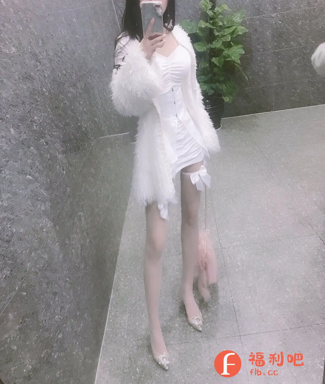 照片中,一位身着白色套装的年轻女子站在一处现代建筑的室内,正在对着镜子自拍。她留着深色长发,手持智能手机,手机屏幕上显示着拍摄界面。她身穿一件白色连衣裙,外搭一件白色毛绒外套,整体造型显得十分甜美可爱。连衣裙设计独特,腰部有类似塑身衣的线条,凸显身材。腿上穿着白色丝袜,丝袜上方系着白色蝴蝶结装饰,更增添了少女感。脚上搭配一双带有水钻装饰的高跟鞋,精致闪亮。旁边还挂着一个粉色的毛绒包包,增加了整体造型的俏皮感。背景是一面灰色的大理石墙面,旁边角落里放置着一盆绿植,为画面增添了一些生机。整体来看,这套服装搭配风格统一,色彩和谐,突显了女性的柔美和时尚品味,可能是一张买家秀照片,用于展示服装的穿着效果。