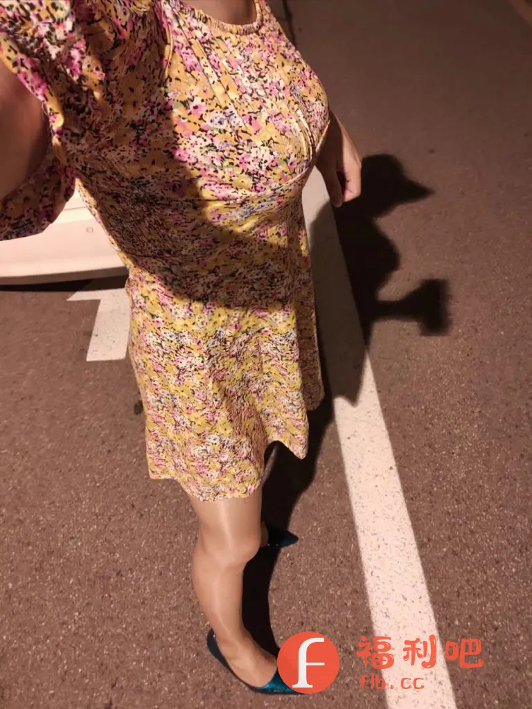 图片展示了一位穿着黄色碎花连衣裙的女士,看起来像是在路边或停车场。连衣裙颜色鲜艳,图案清新,款式简洁大方,很适合春夏季节穿着。女士穿着肉色丝袜和一双深蓝色尖头平底鞋,增添了一丝优雅。拍摄角度是从上往下,可能是在自拍或让朋友拍摄。背景是路面,有一条白色的交通线,光线较暗,可能是在晚上或光线不足的环境下拍摄。整体感觉是时尚休闲,适合日常穿着。
