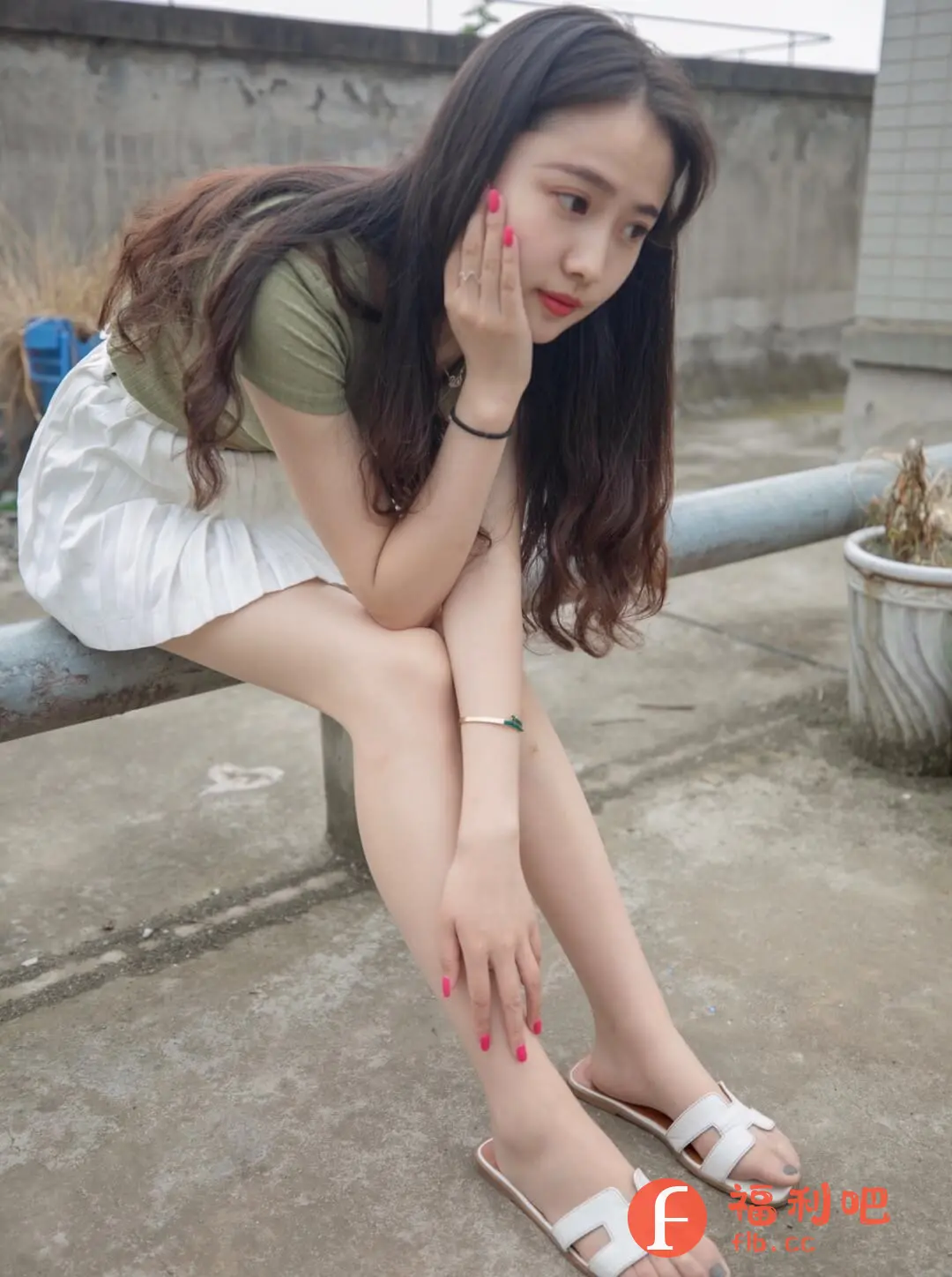 这张图片展示了一位年轻的亚洲女性,她穿着一件军绿色短袖上衣和一条白色百褶短裙,坐在一个灰色的金属栏杆上。她的头发是深棕色,呈现出自然的波浪,自然地垂落在肩上。她一只手托着下巴,另一只手随意地放在腿上,神情略带忧郁,目光看向一边,似乎在沉思。她的指甲涂着鲜艳的粉色。她的手腕上戴着一条细细的手链。她脚上穿着一双白色的凉拖。背景相对简单,可以看到灰色的水泥地面和一个略显斑驳的墙壁。整体风格清新自然,适合用于展示服装、配饰或个人写真。从光线来看,拍摄环境应该是户外,光线较为柔和。