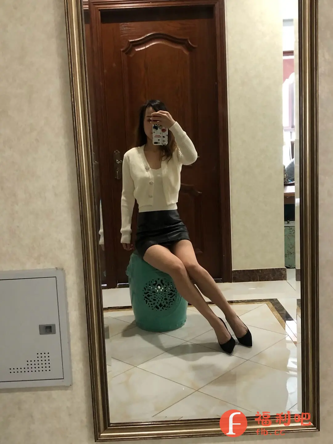 这位美女通过镜子展示了她的时尚穿搭。她身穿一套白色针织衫,内搭同色系背心,下身搭配一条黑色皮裙,脚踩一双黑色高跟鞋,整体造型简约时尚,又不失性感。她坐在一个青绿色的凳子上,腿部线条流畅,更显身材比例。背景是家庭环境,棕色的木门和瓷砖地板,更显生活气息。整体来看,这是一张高质量的买家秀,能够很好地展示服装的款式和上身效果,激发潜在消费者的购买欲望。