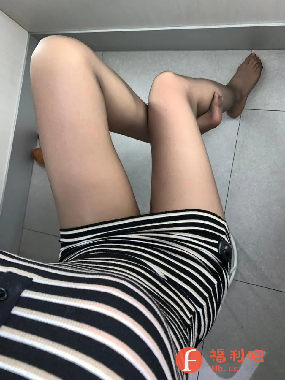 这张照片展示了一位年轻女性的休闲装扮,拍摄视角从上方俯视。她身穿一件黑白条纹相间的连衣裙,条纹方向为横向,勾勒出身体的曲线。裙子的款式简洁,左侧似乎有纽扣设计,整体风格时尚。女性的双腿交叠着,膝盖处微微弯曲,姿态放松。她穿着肉色丝袜,衬托出腿部肌肤的光滑质感。背景是灰色的瓷砖地板,简洁干净。照片整体光线柔和,呈现出一种随性自然的生活状态,适合在电商平台上作为服装展示图。买家秀的真实感更能吸引潜在消费者的目光。