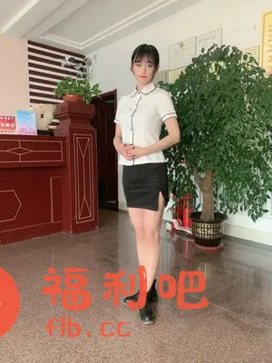 图中一位年轻的亚洲女性站在室内,她身着一套职业装,包括一件白色短袖衬衫和一条黑色包臀裙,脚上穿着一双黑色皮鞋。衬衫的领口和袖口带有黑色滚边,显得精致而干练。裙子的侧边有一个小开叉,为整体造型增添了一丝时尚感。她留着齐刘海,头发扎成简单的发髻,双手交叠放在身前,姿态端庄优雅。背景是一个类似酒店大堂的环境,有一个红棕色的前台,前台上有盆栽作为装饰。旁边有一颗茂盛的盆栽植物,增添了室内的生机。墙上挂着一些证书或奖状,显示出这个场所的专业性。整体的光线明亮,场景整洁,营造出一种专业而舒适的氛围。这位女性的形象与环境相协调,给人一种干练、自信的印象。