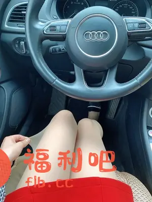 这是一张在车内的自拍照片,拍摄视角是从上往下。画面中心是一名女性,她坐在驾驶座上,双腿并拢,穿着肉色丝袜和红色短裙。她的右手拿着丝袜边缘,似乎在展示丝袜的质感或颜色。背景是汽车的驾驶舱,包括方向盘、仪表盘和中控台。方向盘上清晰可见奥迪的标志。整体画面色彩鲜艳,突出展示了丝袜的细节和穿着效果。这张照片很可能是在展示或推销丝袜产品,旨在吸引潜在买家。构图简洁,重点突出,能够有效展示产品的特点。